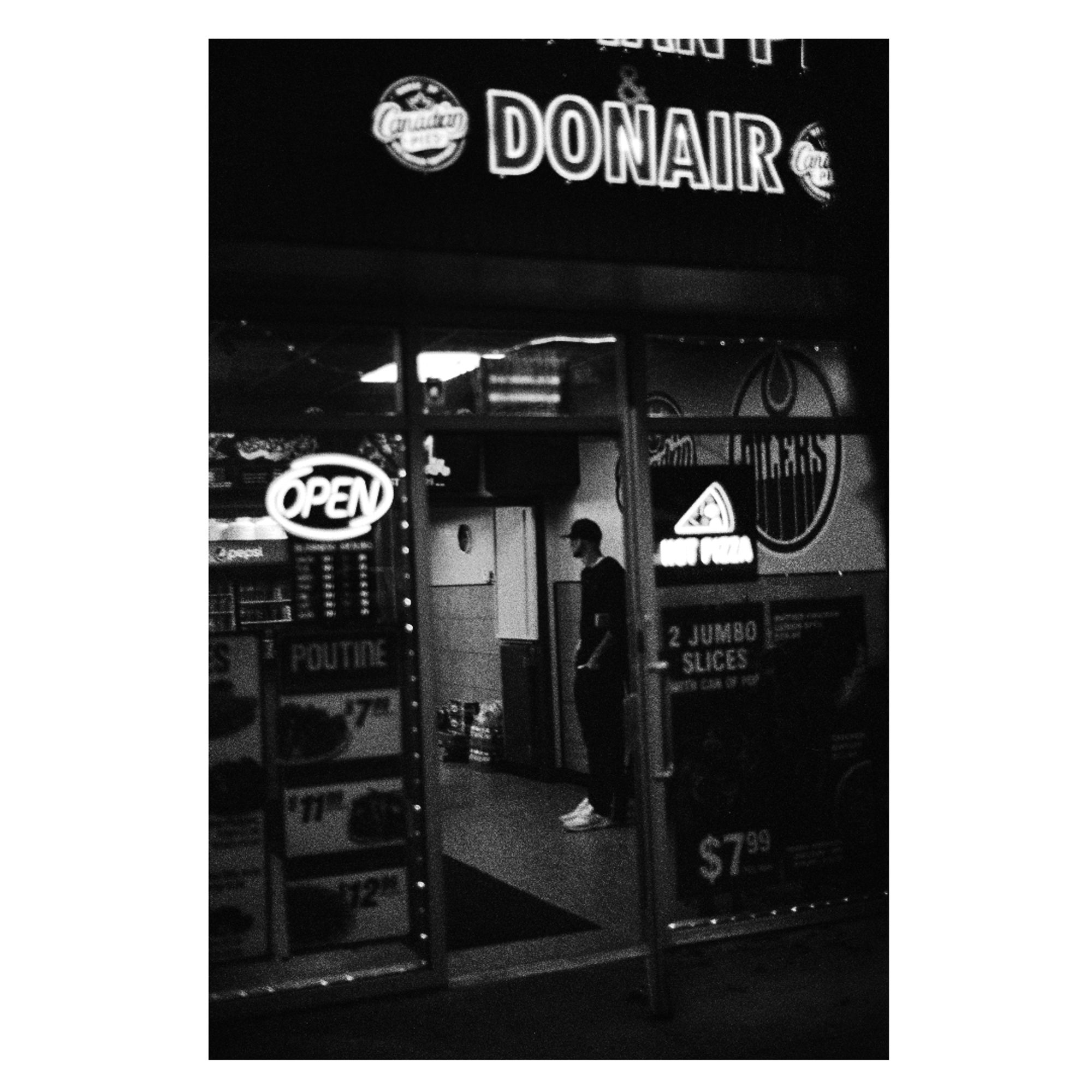 Black and white film photograph in a white border. A man waits for an order at a pizza and donair store. We see him through the open front door. On the front of the business, the open sign and the "DONAIR" of the business name burn bright.

Take October 2024 by Jacy Eberlein in Edmonton Alberta with a Nikon FE 50mm f1.8 E lens and original formula expired AGFA 100.