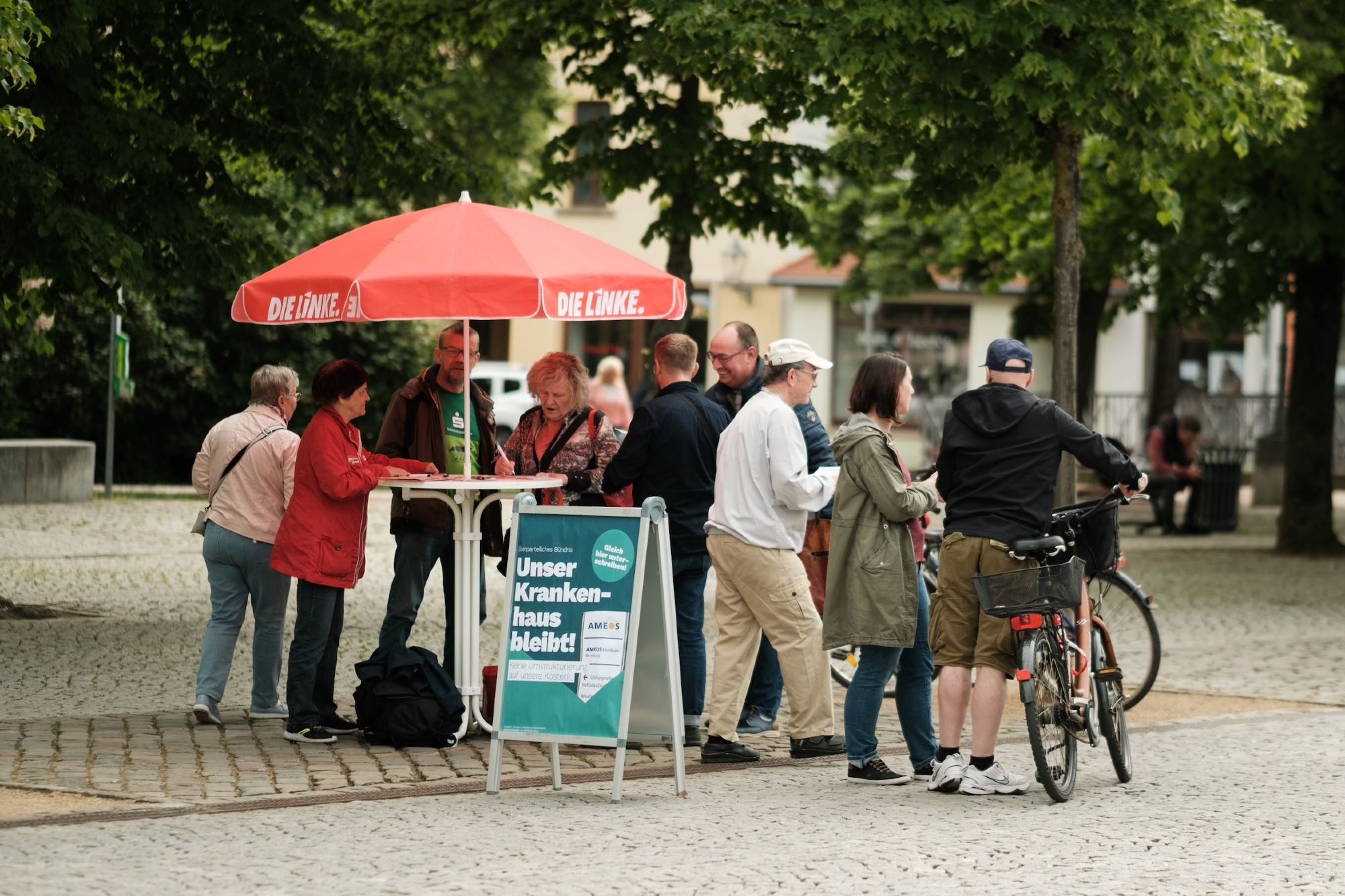 Eine Traube an Menschen steht um einen Infostand mit Stehtisch und rotem Die Linke-Schirm. Davor ein Kundenstopper, der für eine Kampagne zur Rettung des Bernburger Krankenhauses wirbt: „Unser Krankenhaus bleibt!“