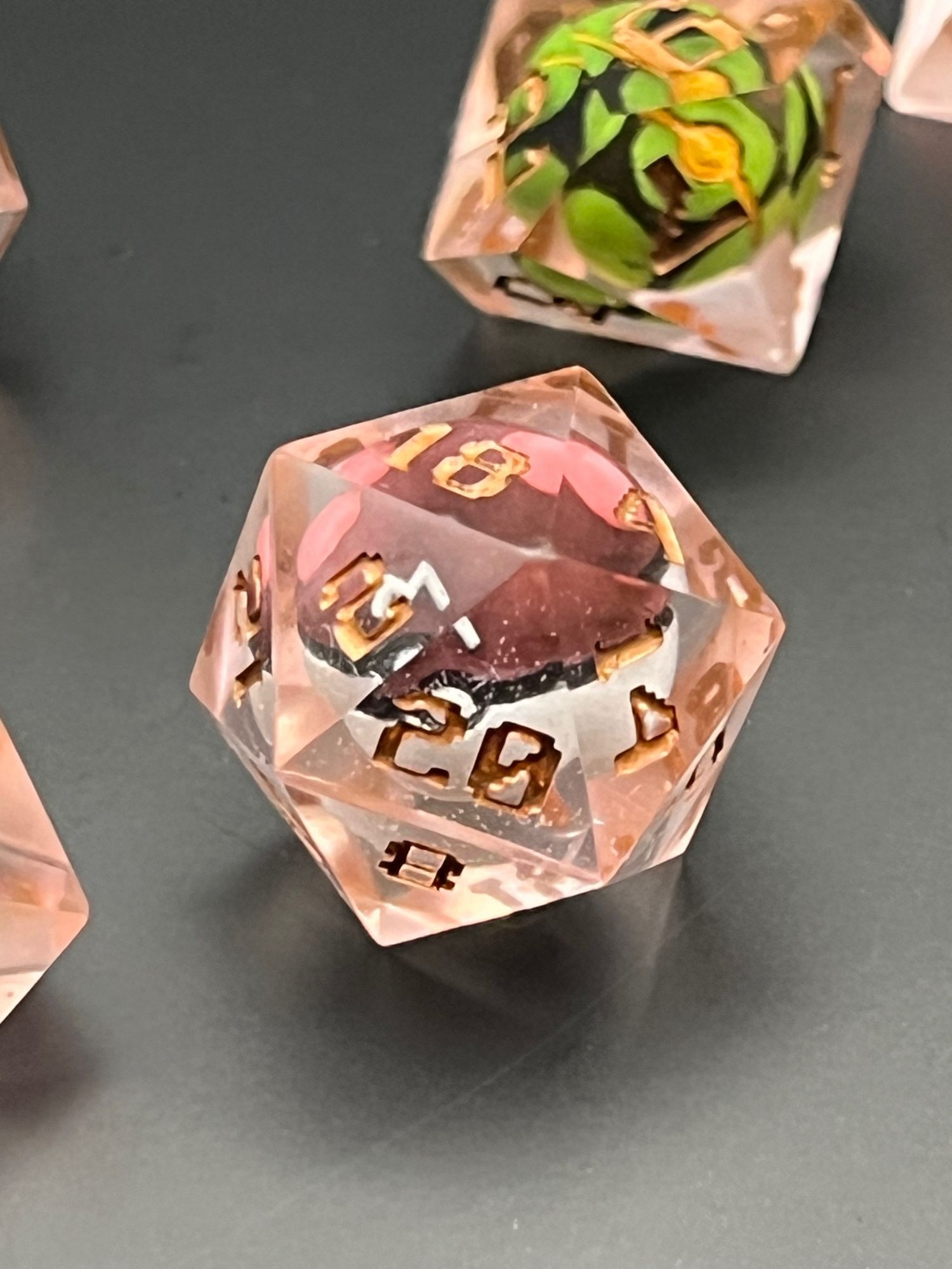 Two rose-gold colored Pokemon ball dice, for Dungeons and Dragons.  The resin used came our pinkish (despite intending to be orange), and the lettering of the numbers is GW’s Scorpion Brass, a deep bronze color.  Each dice has a Pokeball inside.  The d10 has a Dusk Ball with green circles on black.  The d20 is a Master Ball with purple and white, and pink boobs on top, with a white ‘M’.