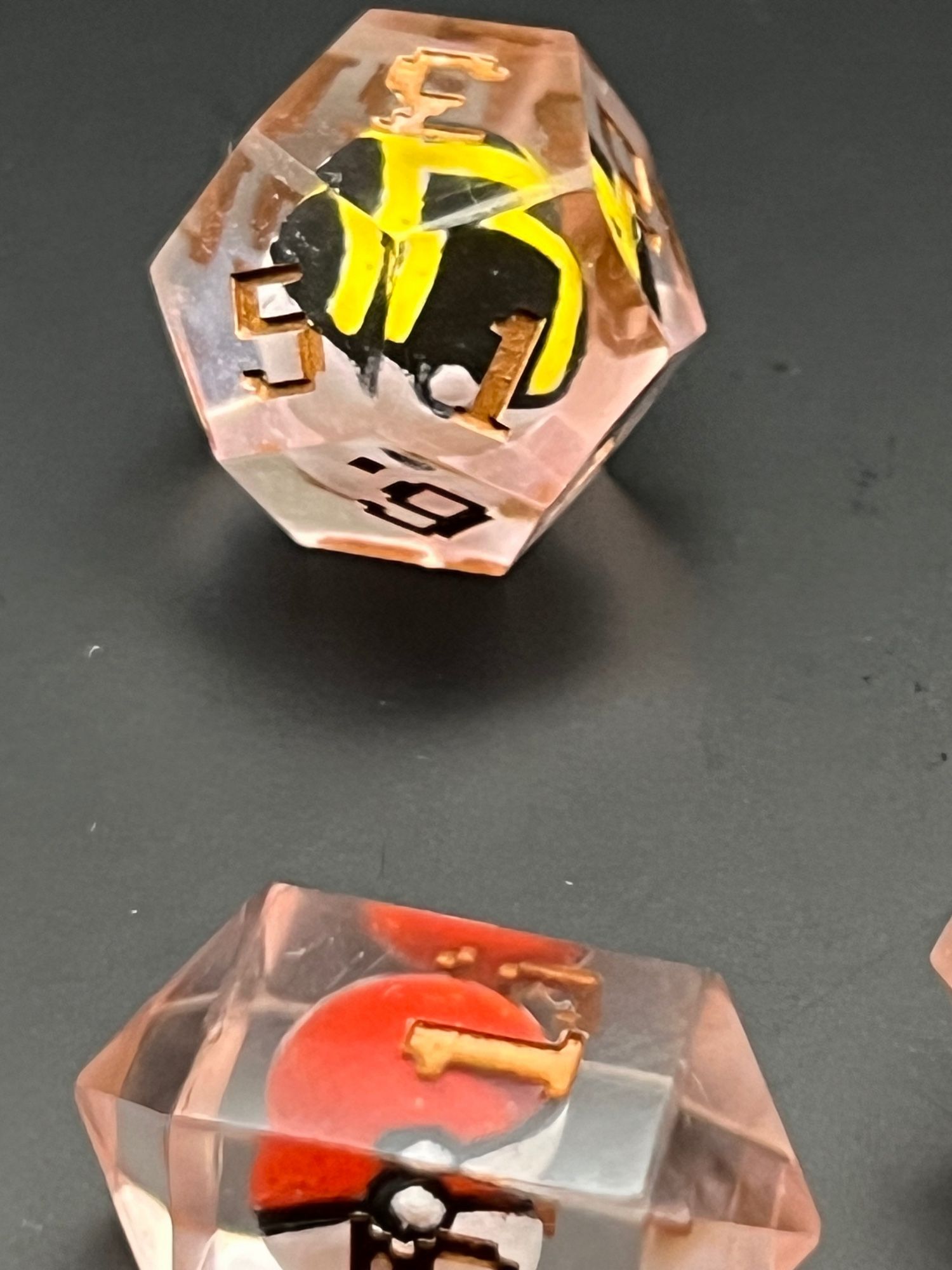 Two rose-gold colored Pokemon ball dice, for Dungeons and Dragons.  The resin used came our pinkish (despite intending to be orange), and the lettering of the numbers is GW’s Scorpion Brass, a deep bronze color.  The d4 has the standard red and white.  The d12 is an Ultimate Ball with black and white and yellow stripes.