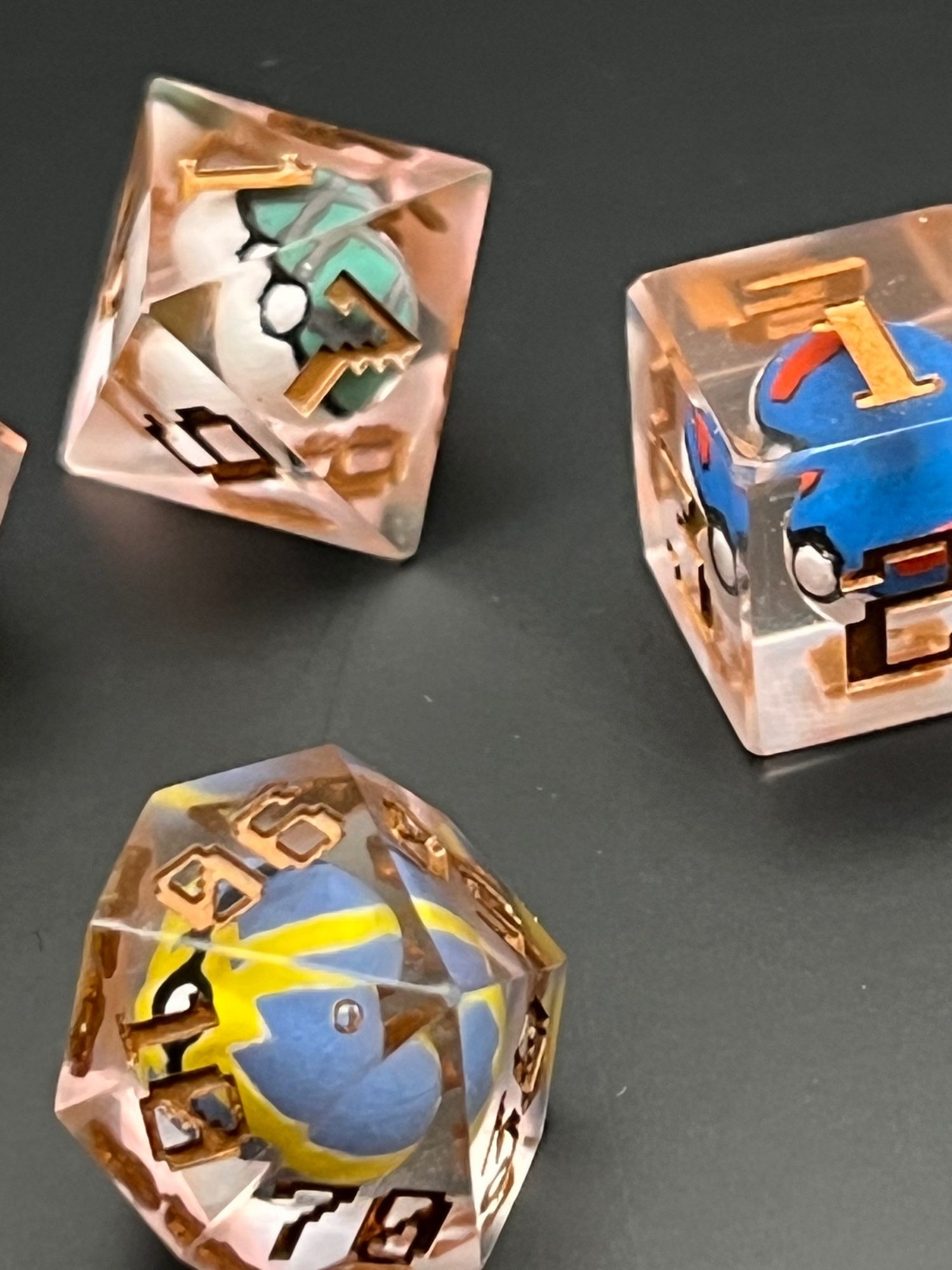 Three 7 rose-gold colored Pokemon ball dice, for Dungeons and Dragons.  The resin used came our pinkish (despite intending to be orange), and the lettering of the numbers is GW’s Scorpion Brass, a deep bronze color.  Each dice has a Pokeball inside. The d6 has a blue and white with red stripes Greatball. The d8 has a Net Ball, with it’s green and white and grey net pattern.  The percentile d10 is a Quick Ball with blue and yellow stripes.