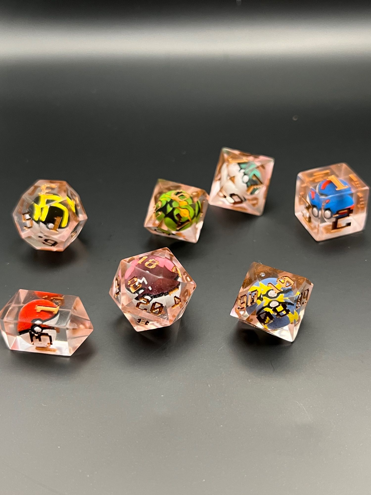 A set of 7 rose-gold colored Pokemon ball dice, with different dice for each that is needed for Dungeons and Dragons.  The resin used came our pinkish (despite intending to be orange), and the lettering of the numbers is GW’s Scorpion Brass, a deep bronze color.  Each dice has a Pokeball inside.  The d4 has the standard red and white.  The d6 has a blue and white with red stripes Greatball. The d8 has a Net Ball, with it’s green and white and grey net pattern.  The d10 has a Dusk Ball with green circles on black.  The percentile d10 is a Quick Ball with blue and yellow stripes.  The d12 is an Ultimate Ball with black and white and yellow stripes.  And the d20 is a Master Ball with purple and white, and pink boobs on top, with a white ‘M’.