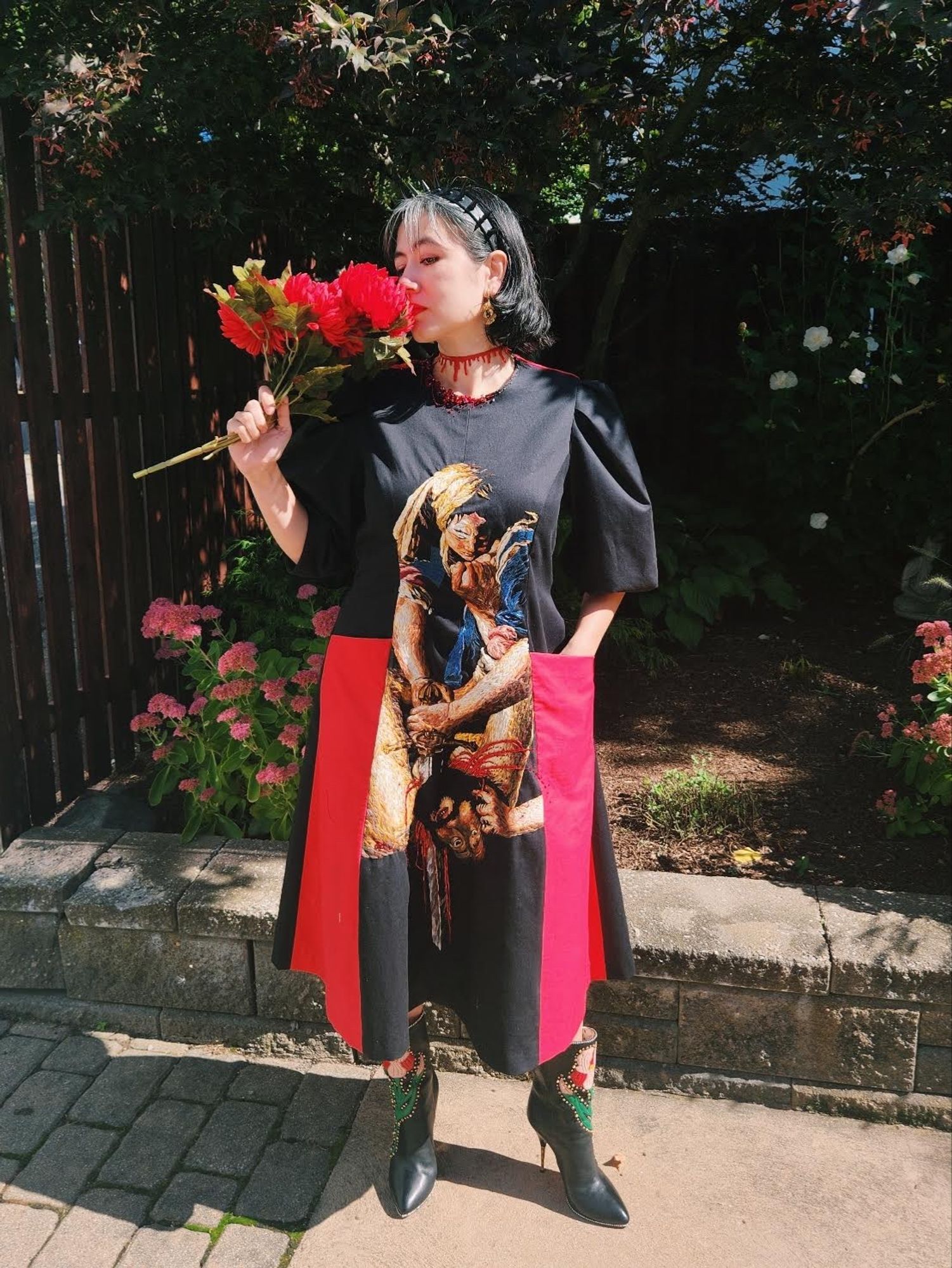 Me pretending to smell fake flowers. I’m wearing a loose fitting black dress with red pockets that go down to the hem. In the center is an embroidery and beadwork recreation of Artemisia Gentileschi’s Judith Slaying Holofernes.