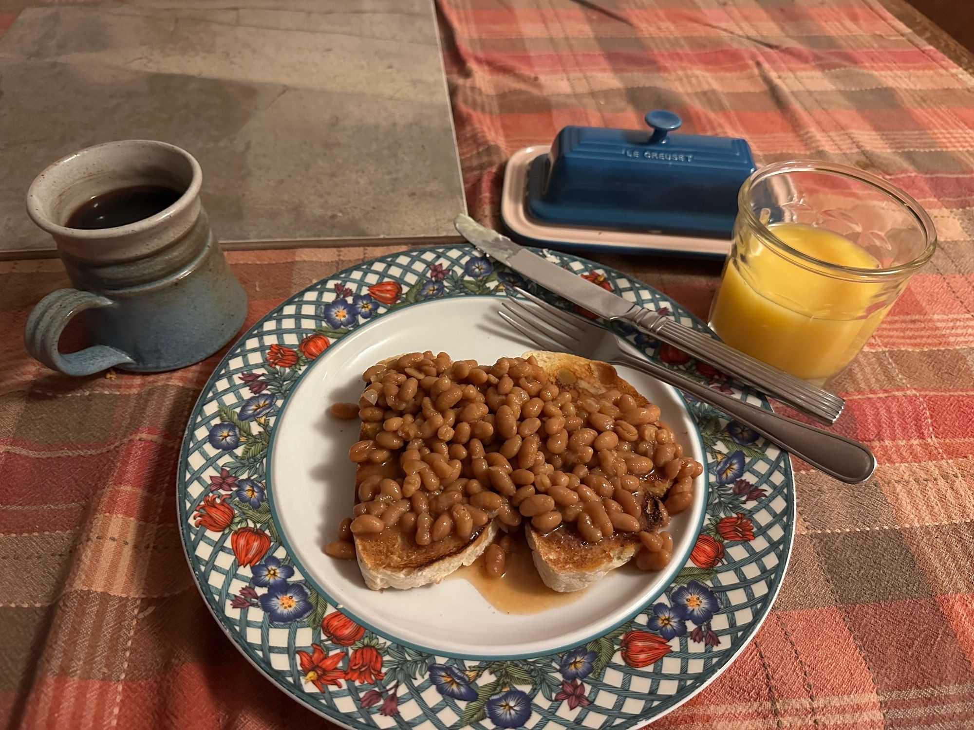 plate with two pieces of sourdough bread toast covered in baked beans with a cup of coffee and a glass of orange juice.  blue Butter dish is on the table above the plate of beans