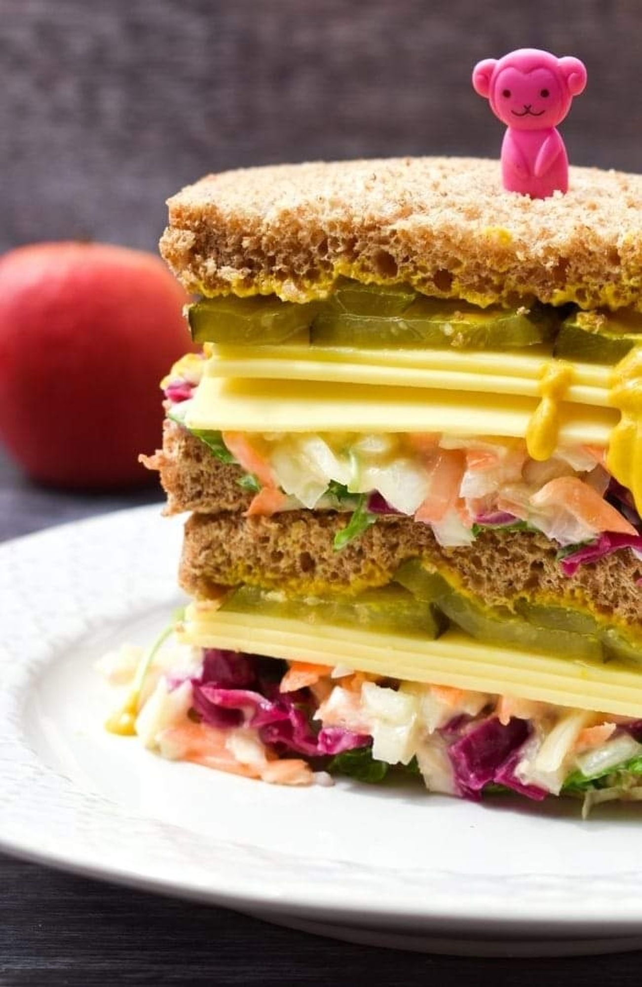 two Deli Sandwiches stacked high on a plate next to a red apple.