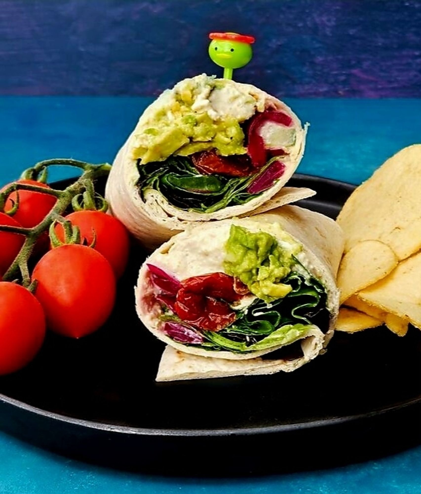 Halved avocado & Hummus wraps on a plate with cherry tomatoes & crisps.