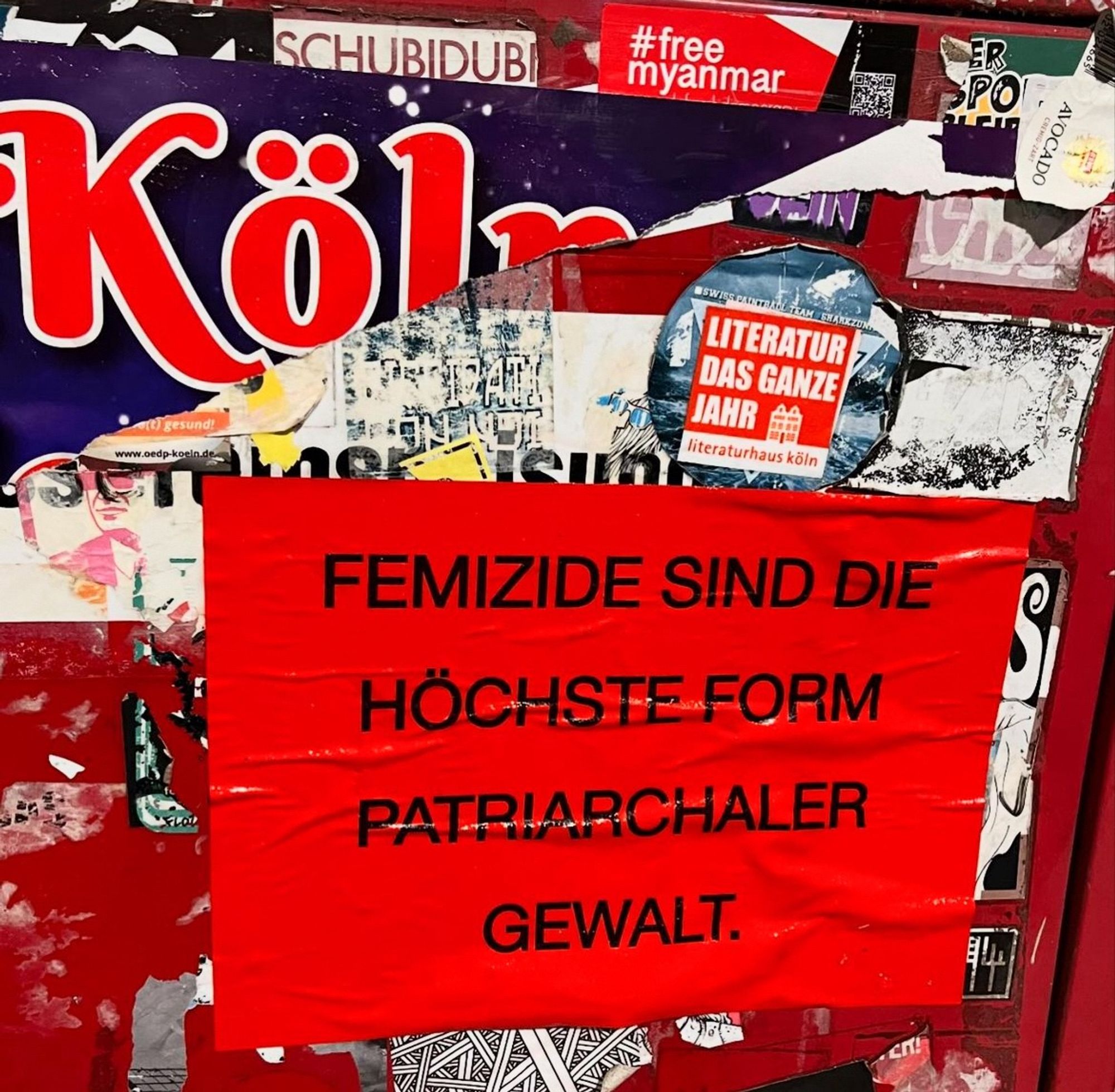 Ausschnitt einer mehrfach überklebten Plakatwand. Oben drauf klebt ein roter Zettel mit der Aufschrift:
Femizide sind die höchste Form patrichaler Gewalt.