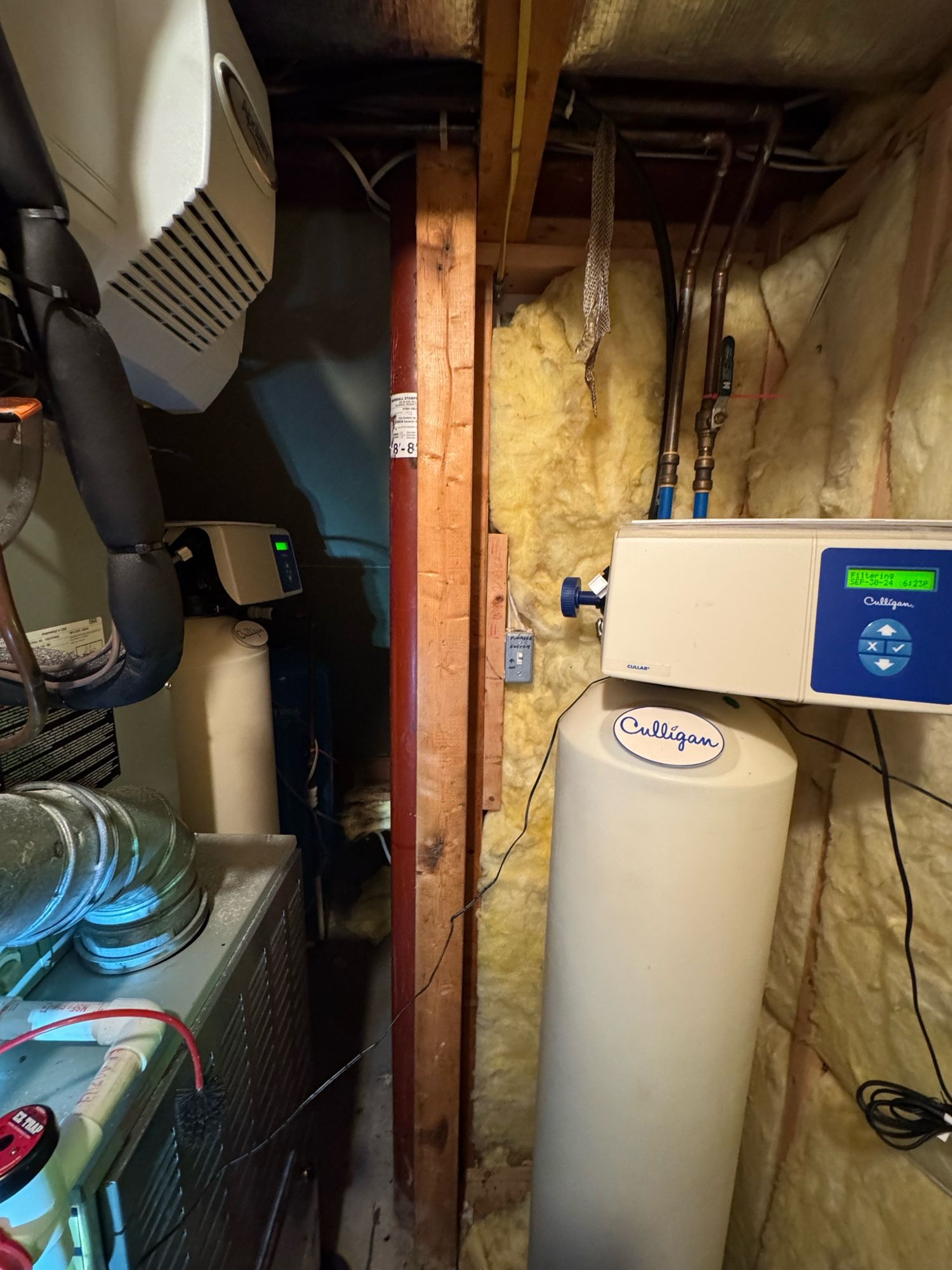 A cramped utility space in a basement showing various water treatment and heating equipment. On the left is a white dehumidifier mounted on the wall. Centered in the image is a large white water softener tank labeled beside another device with digital readings. The area is framed by wooden beams and insulated walls, with visible plumbing and electrical wiring, creating a functional yet crowded setting.