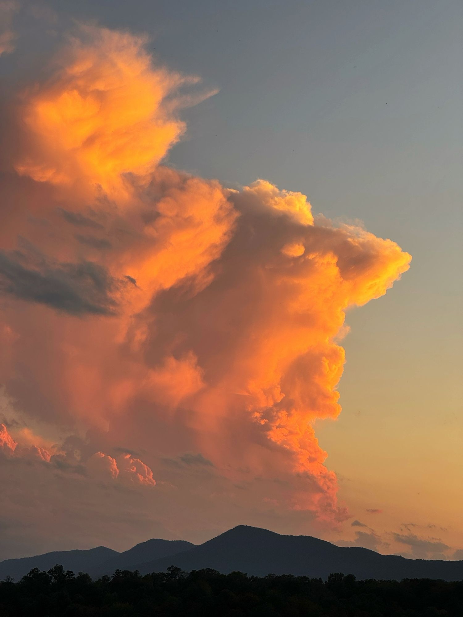 A cloud formation glows with a vibrant orange hue as it catches the last rays of the sunset. The cloud, resembling a fiery plume, rises against a serene sky, fading into a deep blue above and a subdued twilight hue near the horizon. The silhouette of a mountain range in the background adds depth to the scene, emphasizing the cloud’s impressive scale.