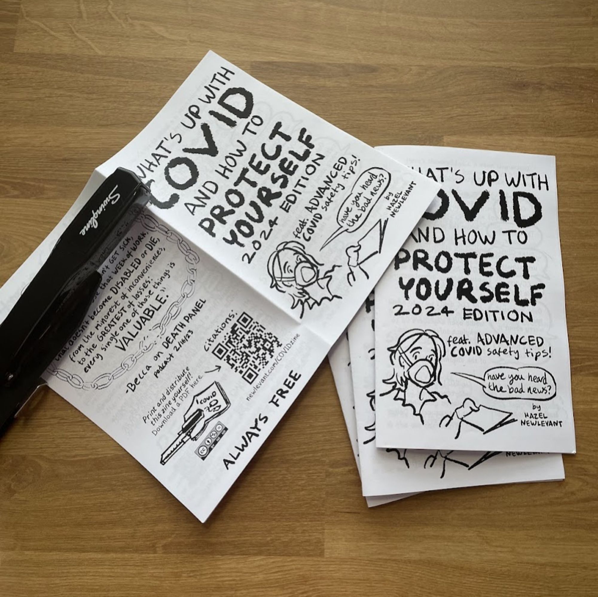 Photograph of a stack of "What's up with COVID" zines, with the top one inside a stapler and about to be stapled.