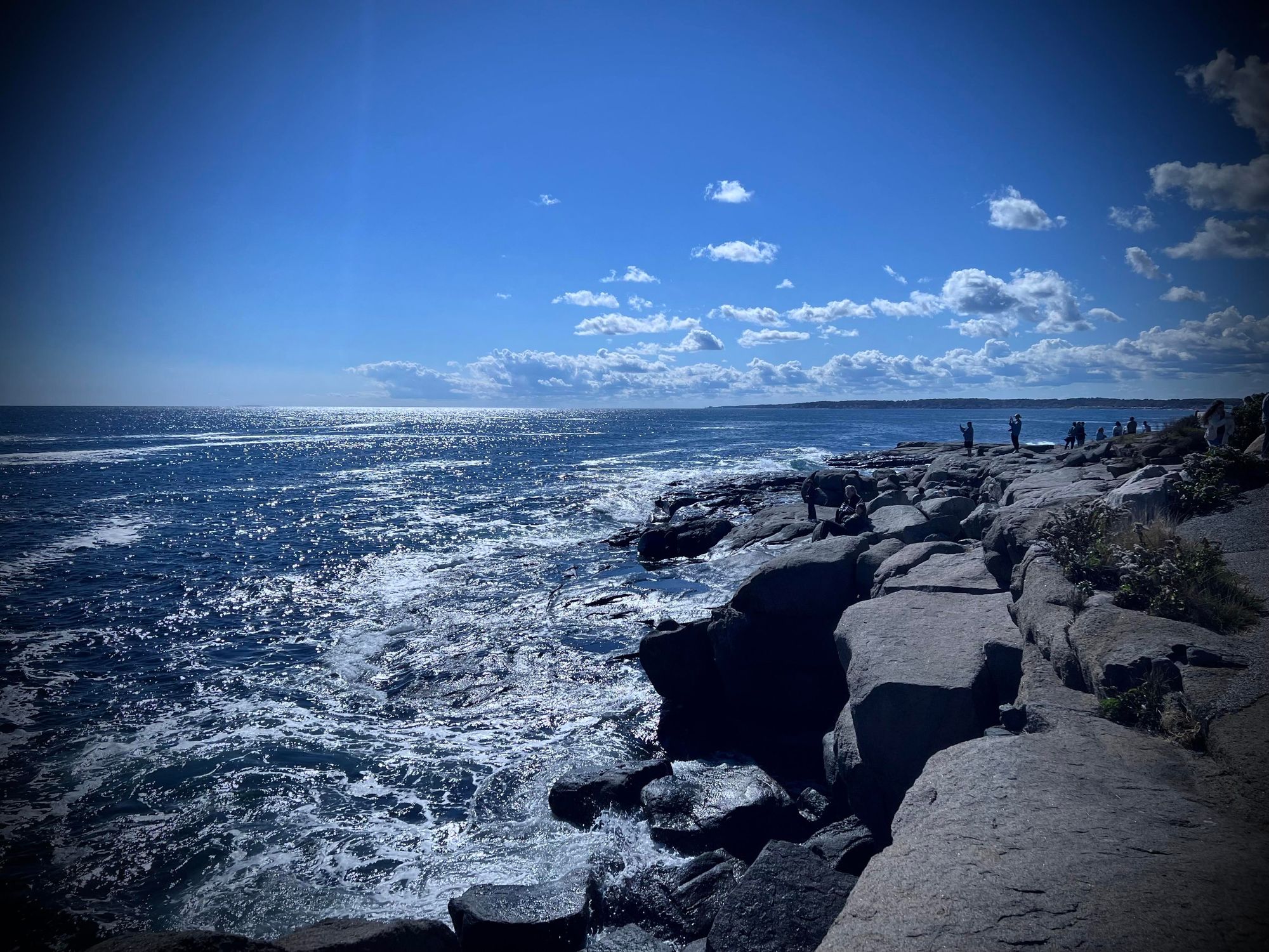 A rugged coastal scene featuring large gray rocks by the ocean, with waves crashing against the shore. The deep blue sky is clear, reflecting the sunlight on the sparkling water.