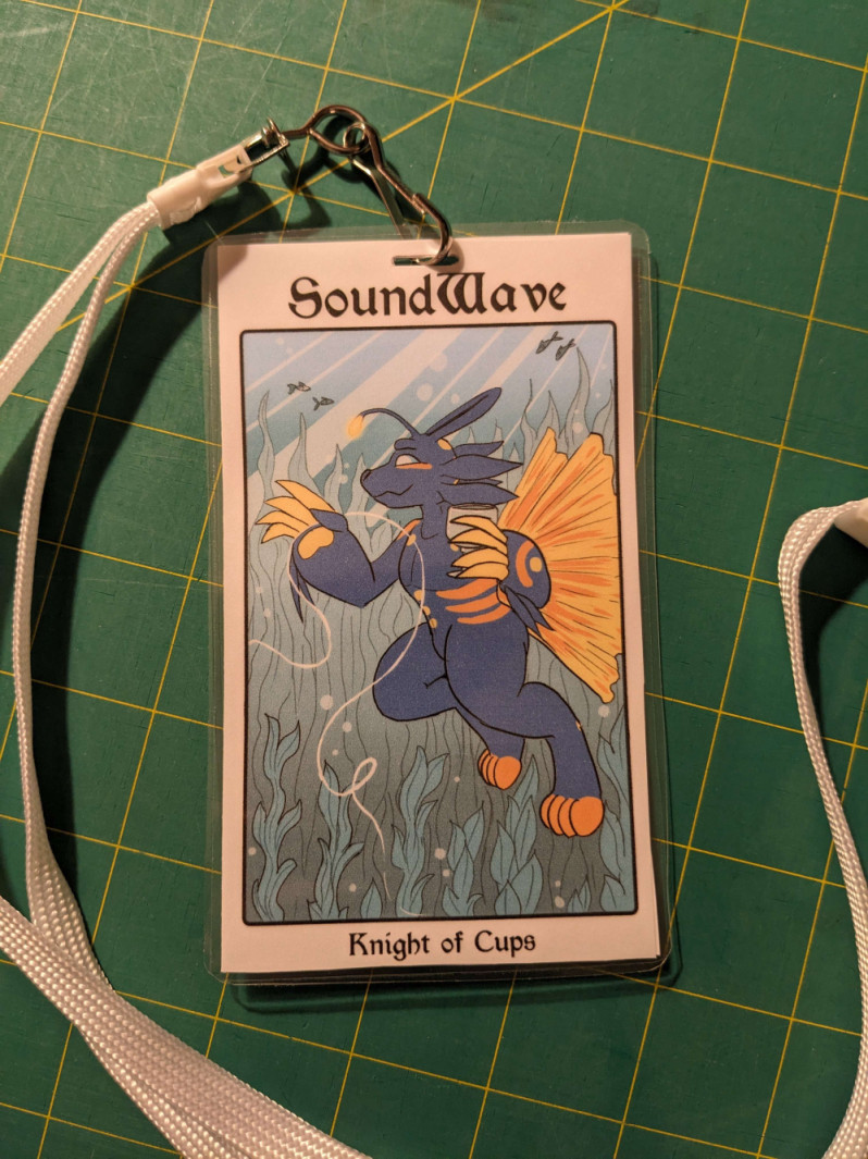 A photograph of a tarot-card themed badge of an anthro fish character, as the Knight of Cups