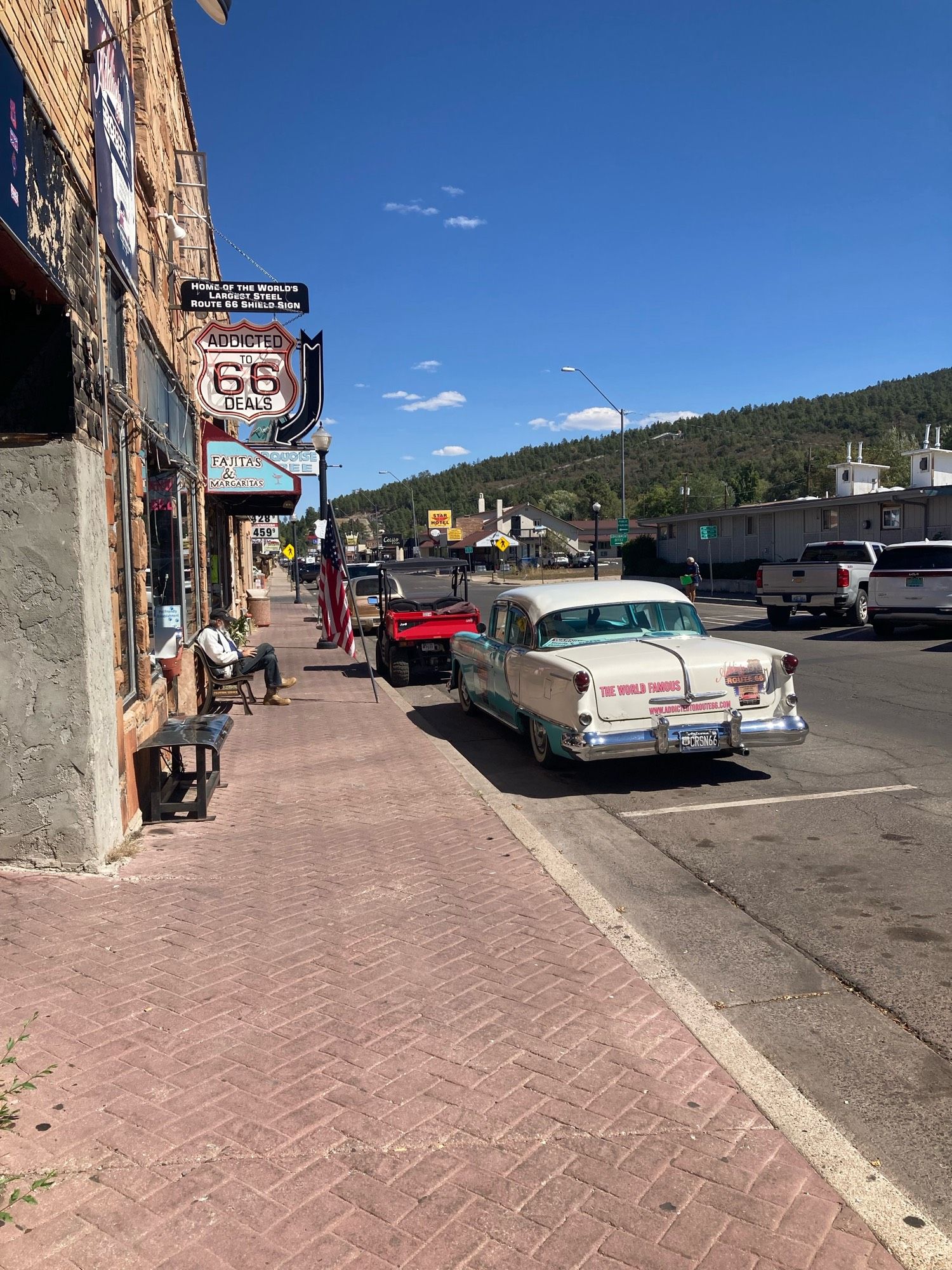 An old Main Street with brick herringbone sidewalk. A vintage white American car is parked in front of a Route 66 store sign. An old man with a cane sits on a bench in front of the store.