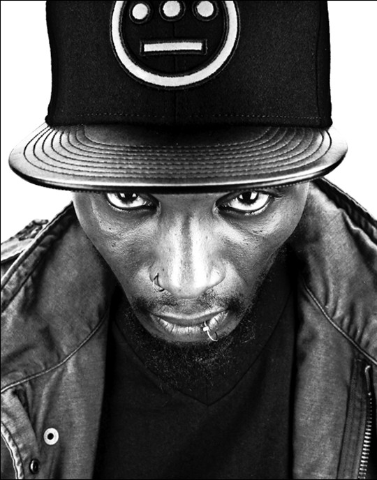 Del the Funky Homosapien is a Black man with a short goatee, a nose ring and lip ring.  He wears a black flat bill cap with a white Hiero logo consisting of a circle with 3 horizontal dots inside and a straight horizontal line under the dots, a black t-shirt and black jacket.  Black and white photograph.