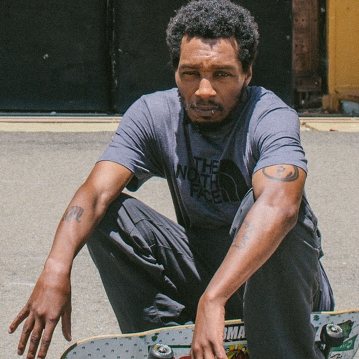 Del the Funky Homosapien is a Black man with short hair, a short beard, a lip ring and tattoos on his arms.  He wears a gray short sleeve t-shirt that says The North Face in black and gray pants.  He squats down on pavement with a freestyle skateboard underneath his left leg.