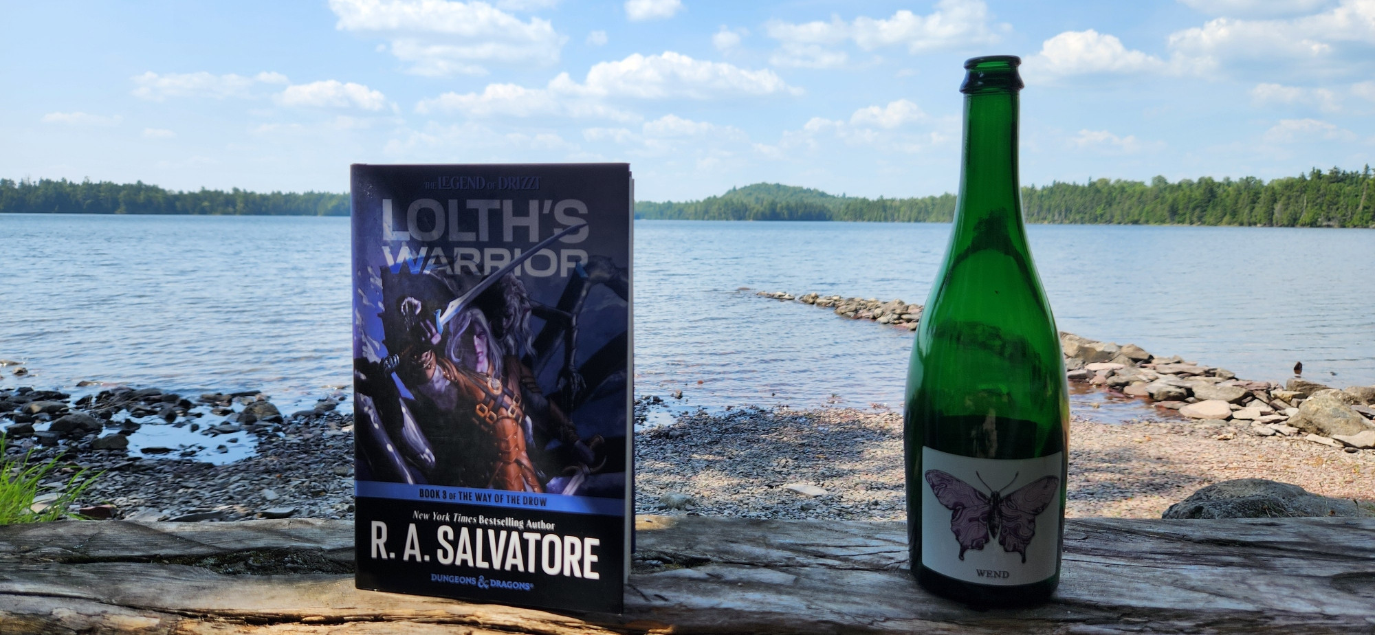 A book titled Lolths Warrior next to a green bottle with a purple butterfly label. In the back ground is a pond with a forested shore and tall hill further away. The sky has patchy white clouds.