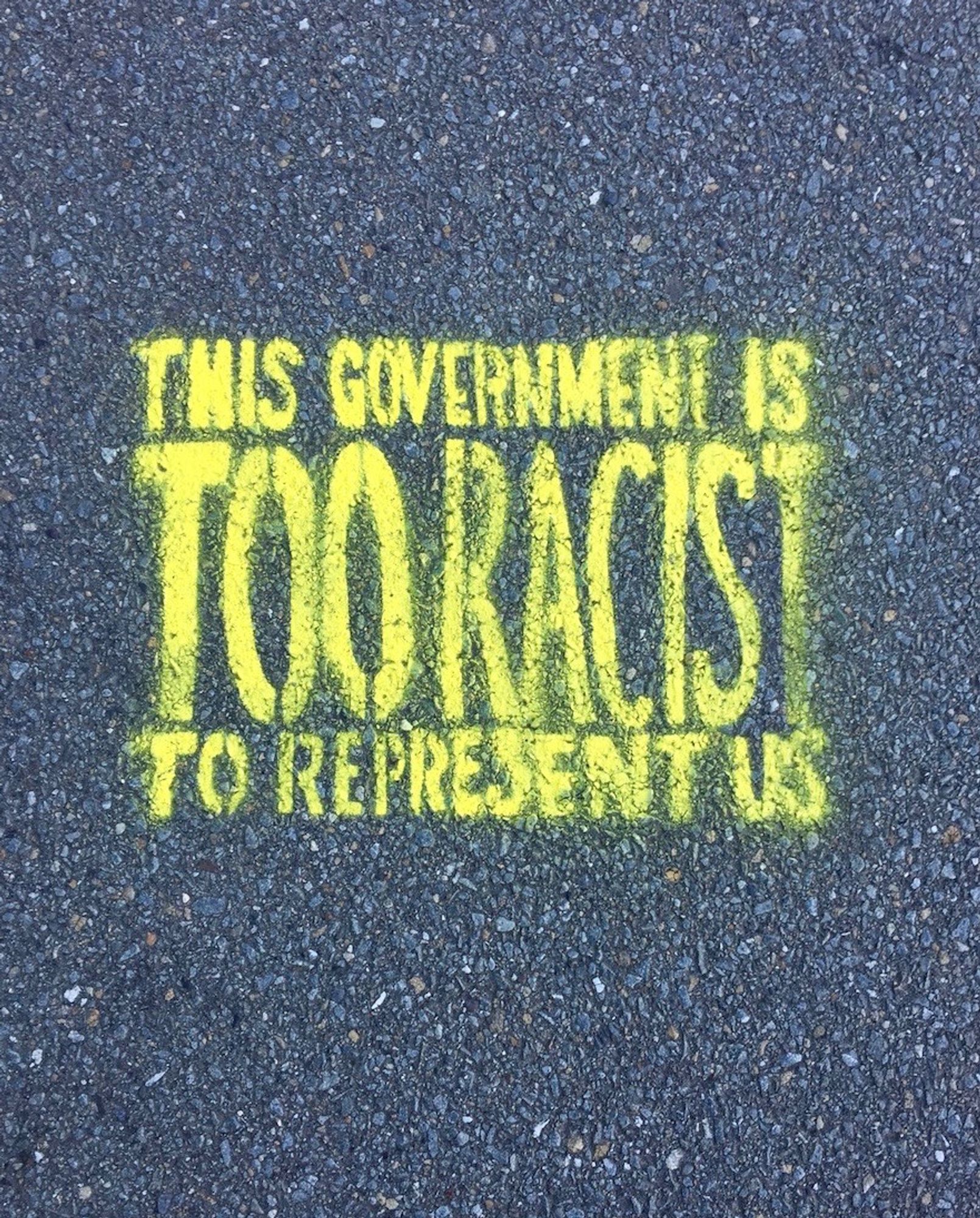 shaded grey footpath with a bright yellow message spray painted on it saying 'THIS GOVERNMENT IS TOO RACIST TO REPRESENT US'