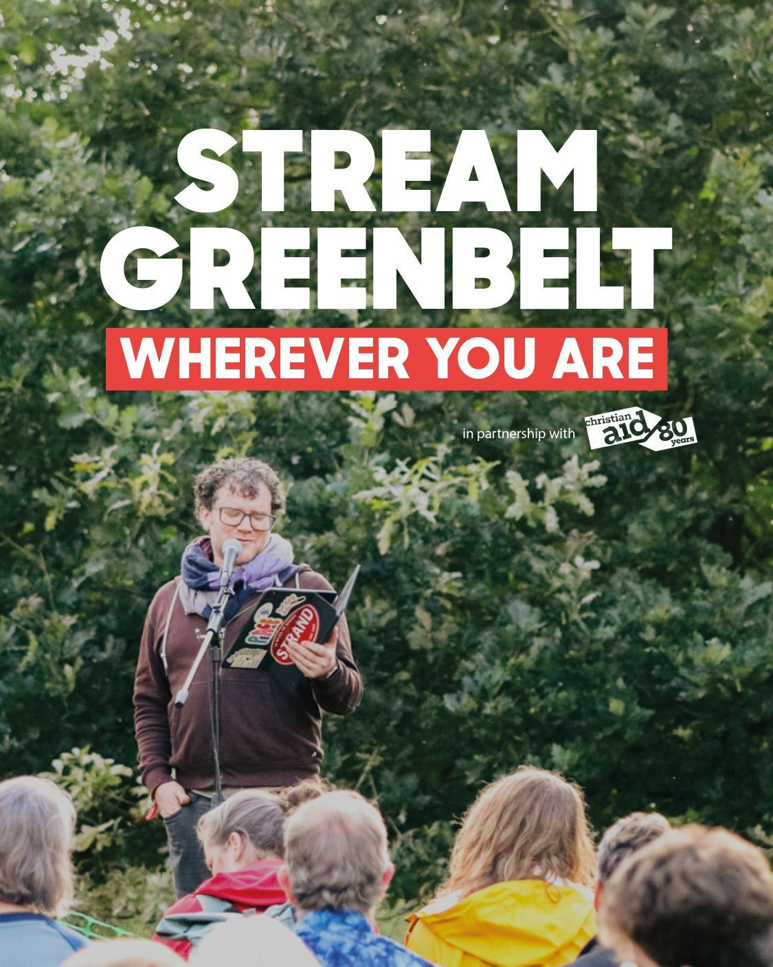 A speaker holds a book while addressing an attentive audience amidst lush greenery, promoting the "Stream Greenbelt" initiative.