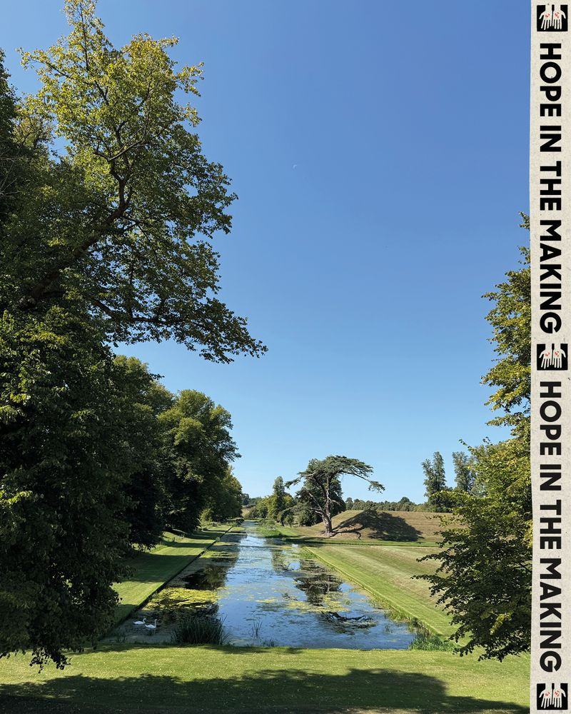 A serene landscape featuring a tree-lined canal reflecting the blue sky, with a lone swan gliding on the water's surface.