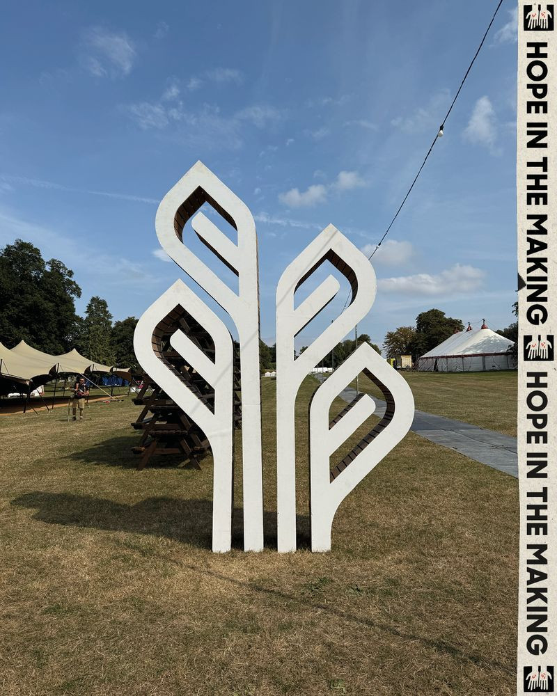 A large, abstract white sculpture resembling stylised leaves stands on grassy grounds, with tents and a circus-style structure in the background.