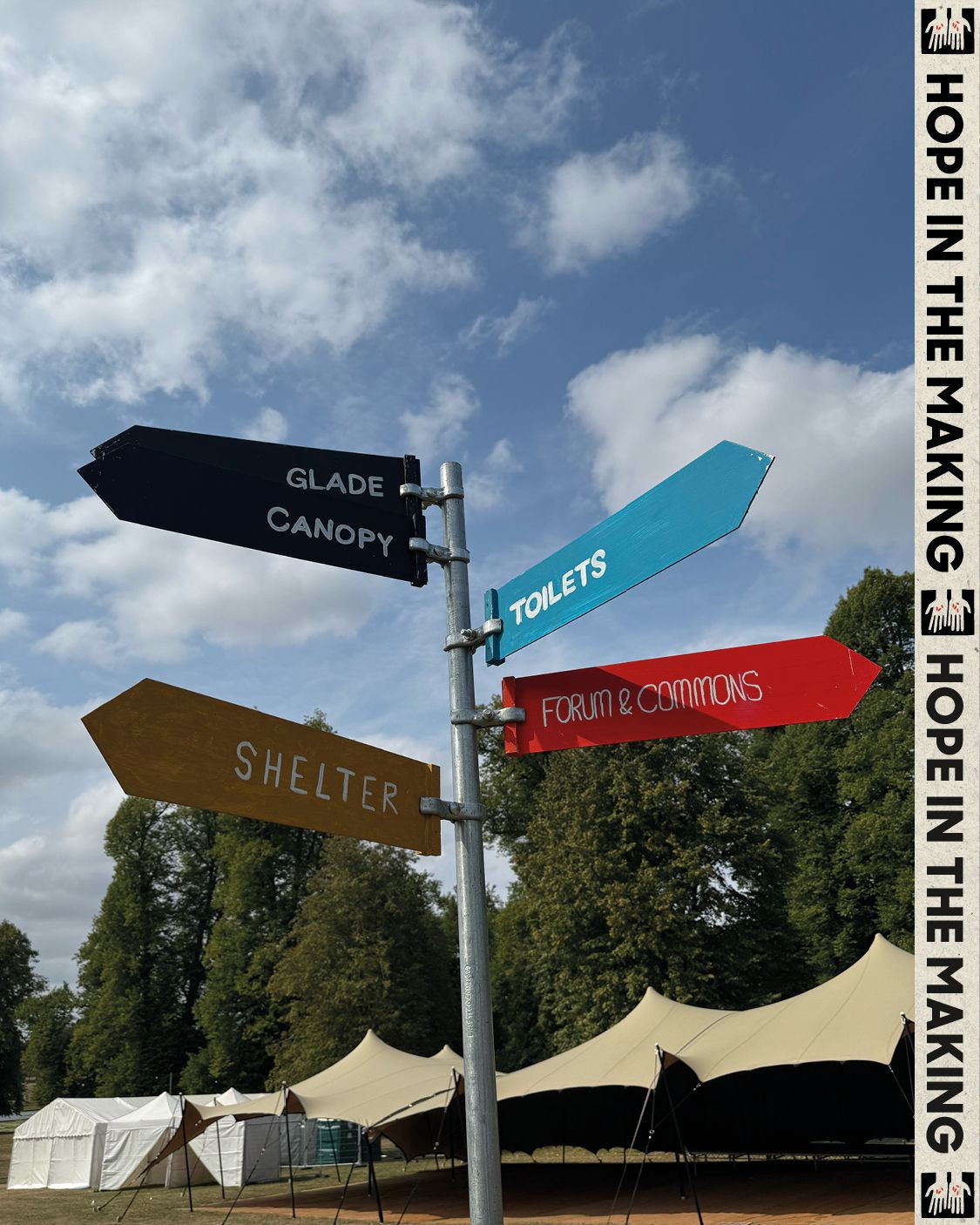 A directional signpost with colourful arrows pointing to 'Glade', 'Canopy', 'Toilets', 'Shelter', and 'Forum & Commons' in a tented area.