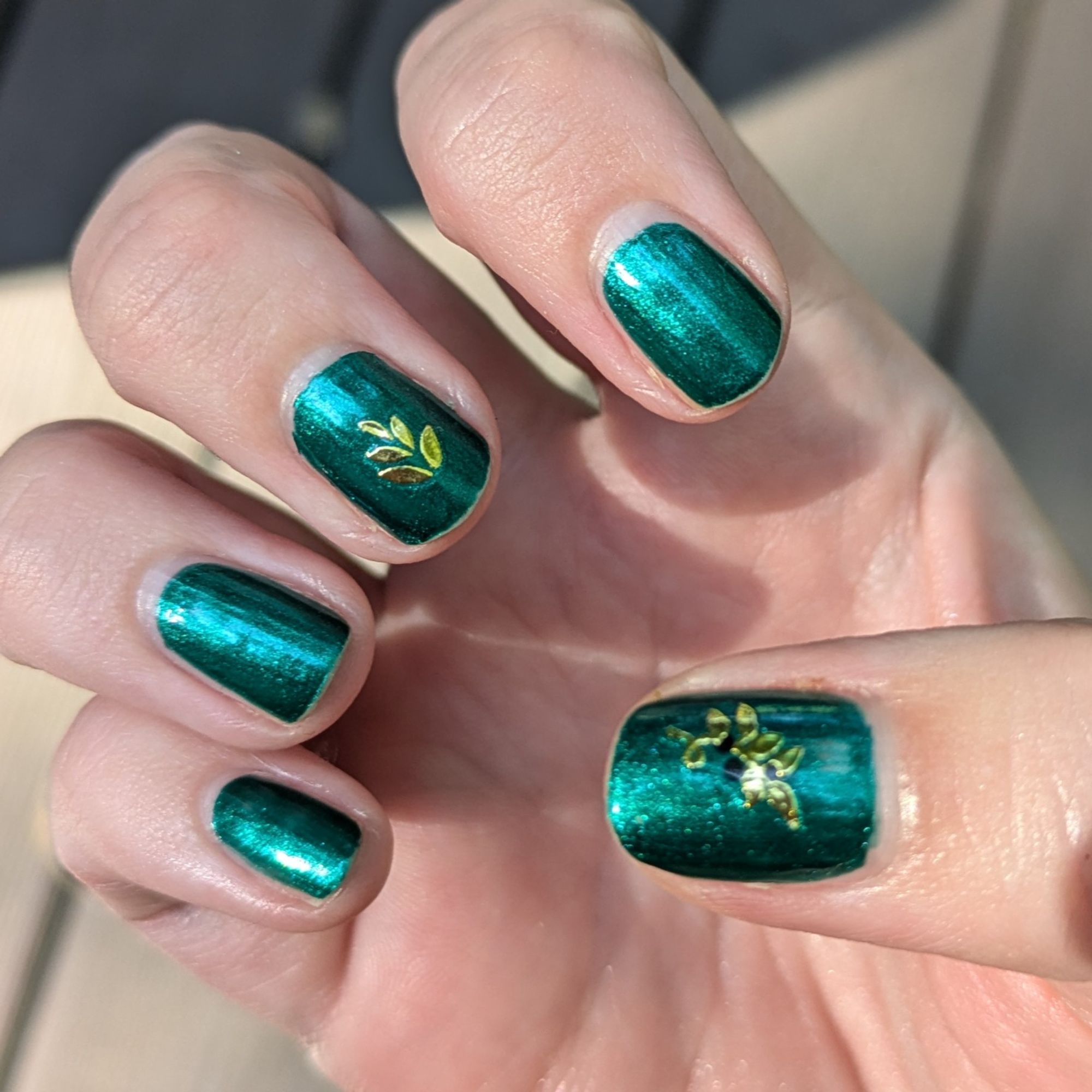 Dark green nails with shiny gold leaf patterns on the thumb and middle nails