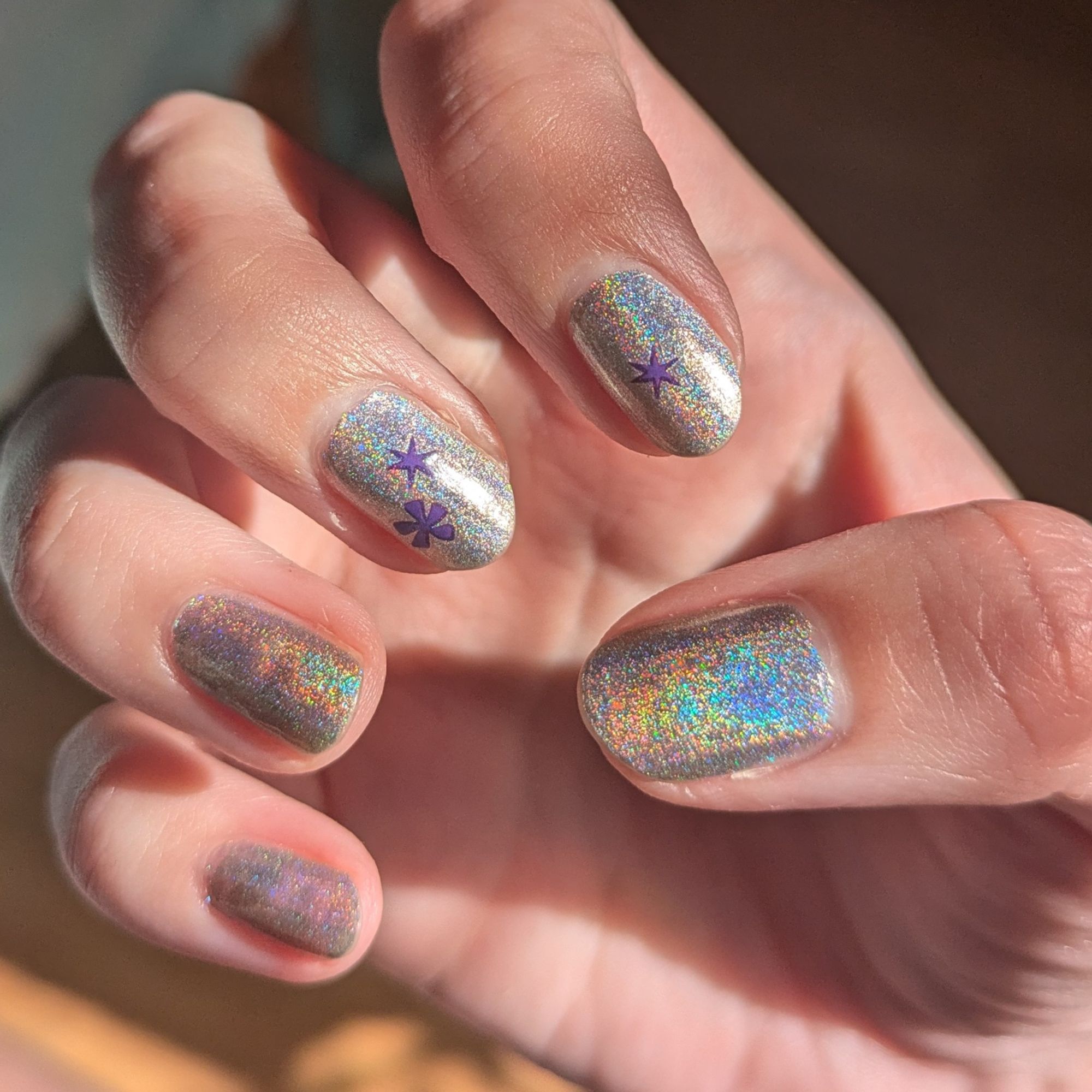 A hand, nails facing the camera. The nail polish is holographic with little purple stars on the index and middle nails.