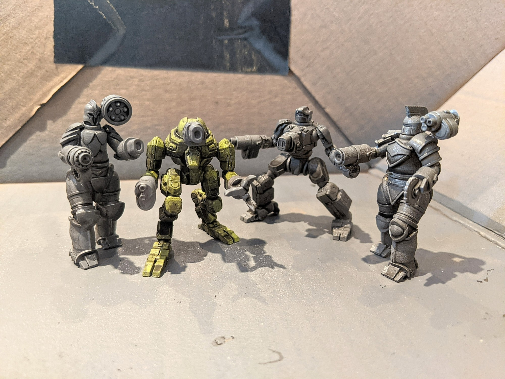 The Titans, left to right, Tethys, Styxx, Eos, and Rhea, in their secondary configurations. They are all grey except for Styxx who is green. They are in a cardboard box with some tape on the wall of the box. The Titans were sculpted by Jason Miller. 