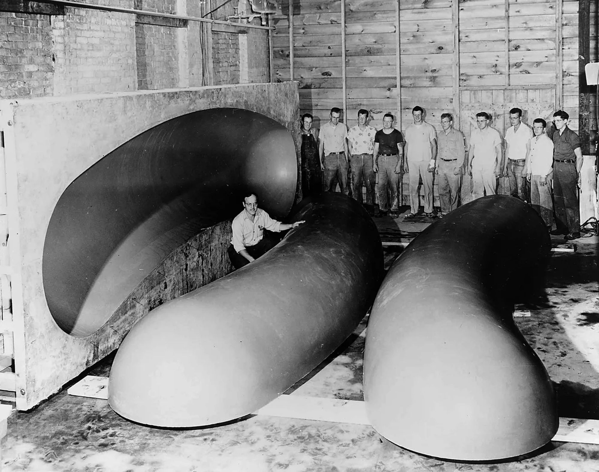 A black and white photo of the fiberglass shell of the OscarMayer Weinermobile next to a mold, and some standard issue mid century white dudes standing in the background