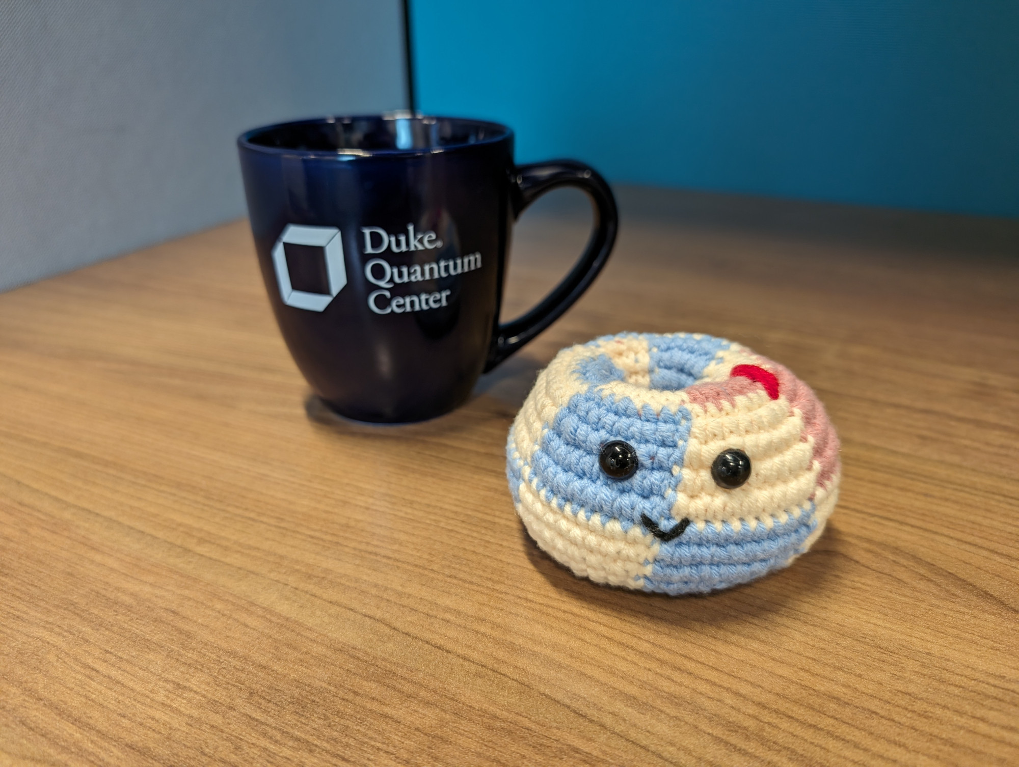 Duke quantum center mug next to a crocheted version of the toric code with one X error, two eyes, and a smile.