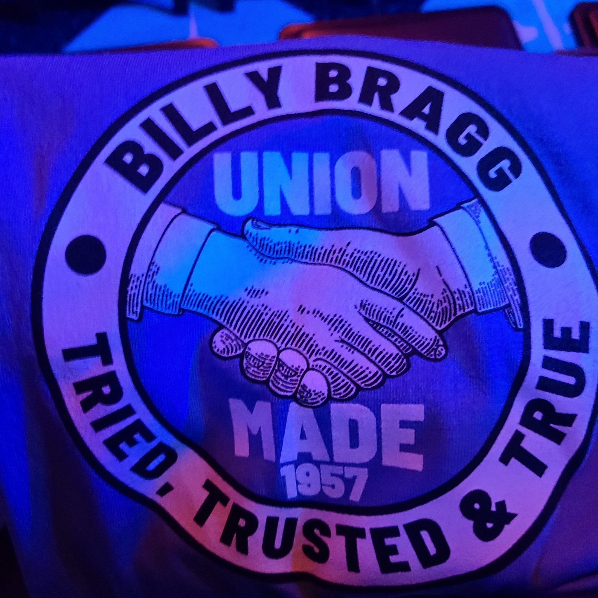 A folded blue t-shirt with a round graphic that says Billy Bragg - Tried, Trusted & True. in the centre of the circle is an image of two hands clasped in a handshake & the words UNION MADE 1957]