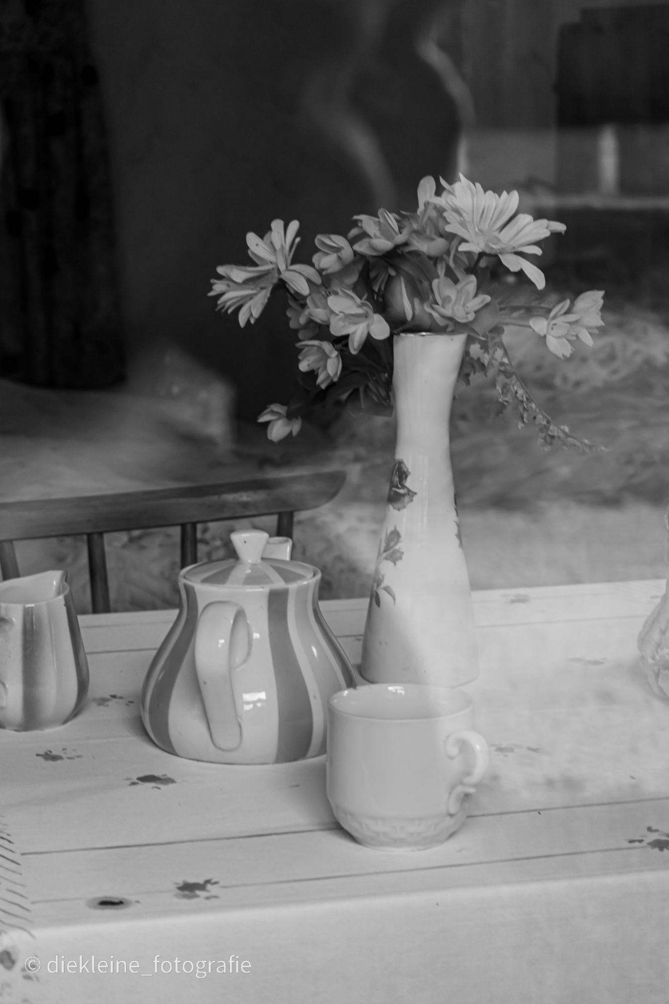 Ein gedeckter Tisch mit einer gestreiften Kanne und einen ebenso gestreiften Milchkännchen. Davor eine weiße Tasse. In der Mitte steht eine Vase mit Blumen drin. De tisch ist durch ein Fenster hindurch fotografiert so dass es eine leichte Spiegelung durch Bild gibt. Im Hintergrund ist eine Stuhllehne zu erkennen .