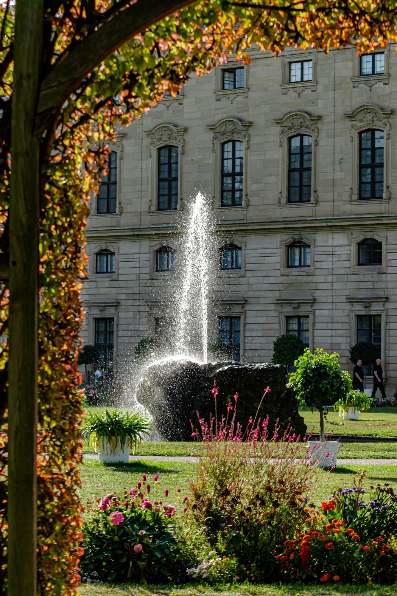 Der Blick geht durch ein Bogen mit Blättern hindurch auf einen Brunnen und eine Hauswand im Hintergrund durch. Vorne dran grüne Wiese mit einigen Blumen und Sträuchern. Der Brunnen ist aus Stein und es sprüht eine Wasserfontänen im hohen Bogen nach links raus.