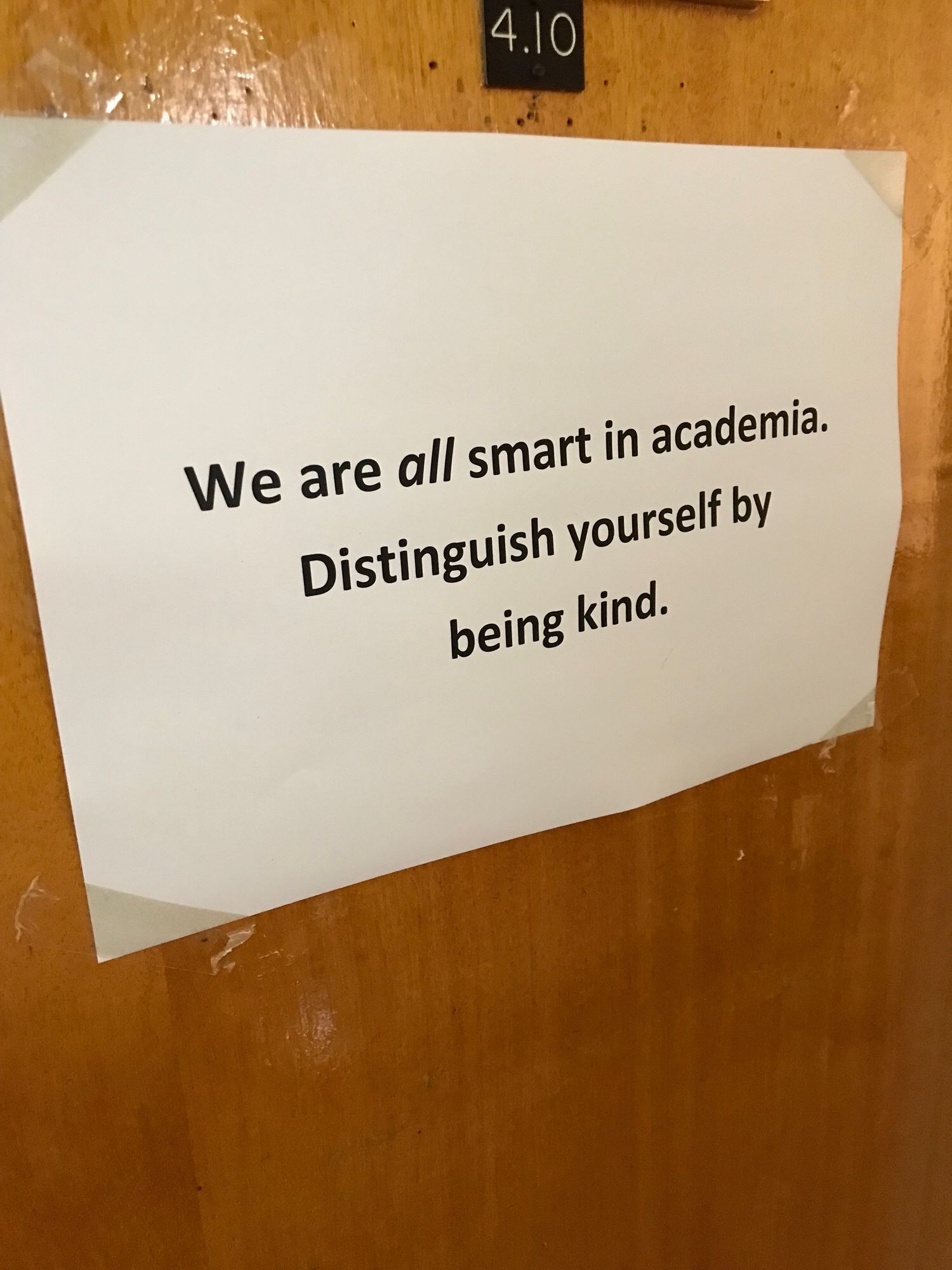 A piece of white A4 paper taped on a brown door. The black text reads: We are ALL smart in academia. Distinguish yourself by being kind.