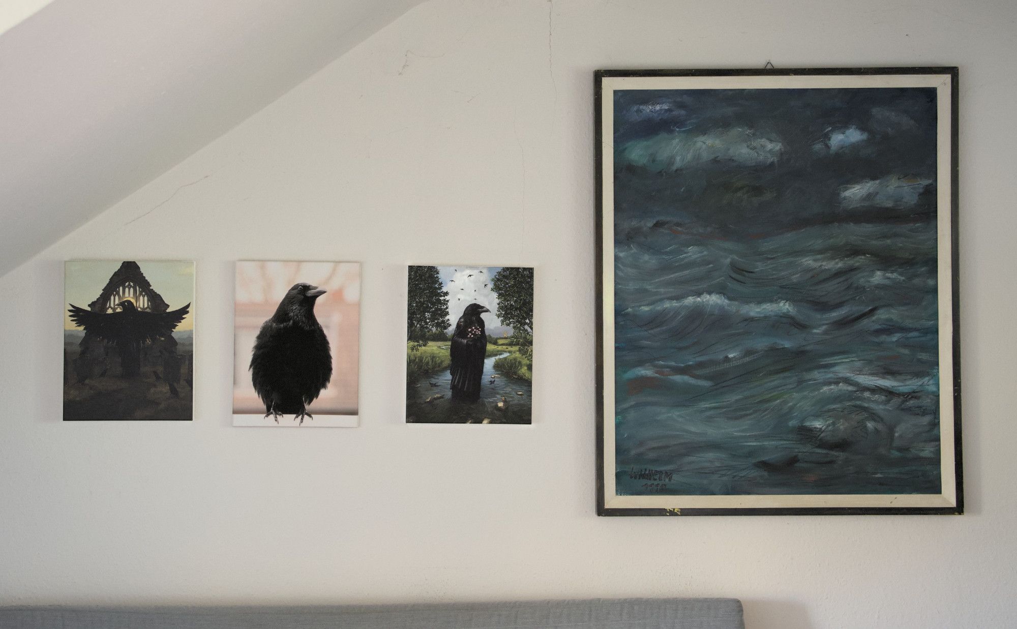 Three paintings on a wall; left to right::

A print of a surreal painting "The gilded teacher and their flock" by Holly Humphries: An anthropomorphic crow with streched out wings reading from a book to their class of jackdaws.

A canvas print of a portrait I took of my best crow friend Kräkrä; Kräkrä is sitting on a window frame, puffed up comfortably, slightly tilted head, looking slightly right, giving him an appearance of a smirky smile.

A print of a surreal painting "The Herald of Summer" by Holly Humphries: An anthropomorphic crow standing in a creek surrounded by meadows in blossom, holding flowers, a butterfly sitting on their right shoulder. Swallows flowing overhead and close to the creek's surface.

An impressionistic painting (original) by Hans Willhelm, a sea of waves under a stormy sky in a dark aquamarine, gray and black color palette.