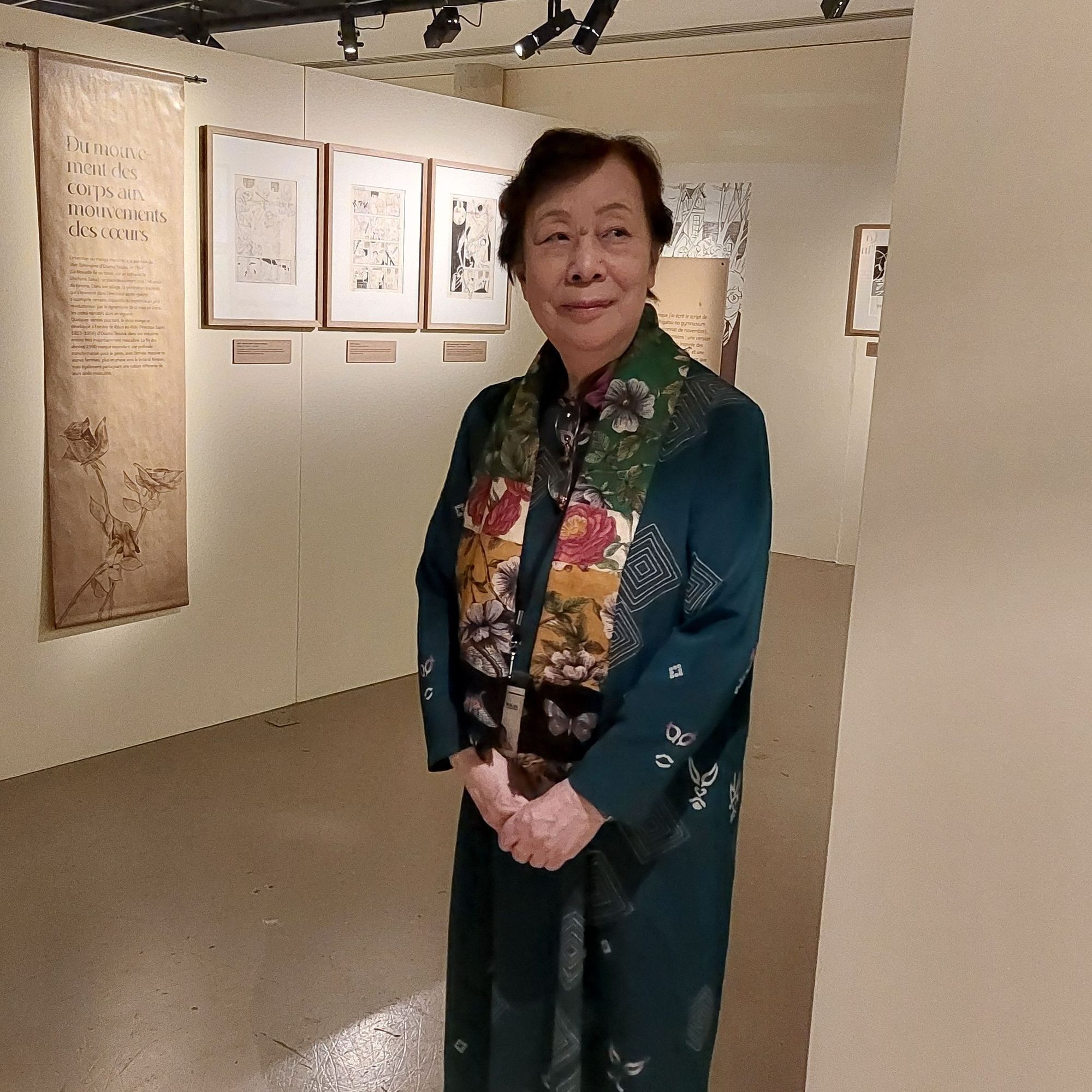 Une femme japonaise se tient les mains, avec un petit sourire. Elle porte une robe vert émeraude et une écharpe fleurie.