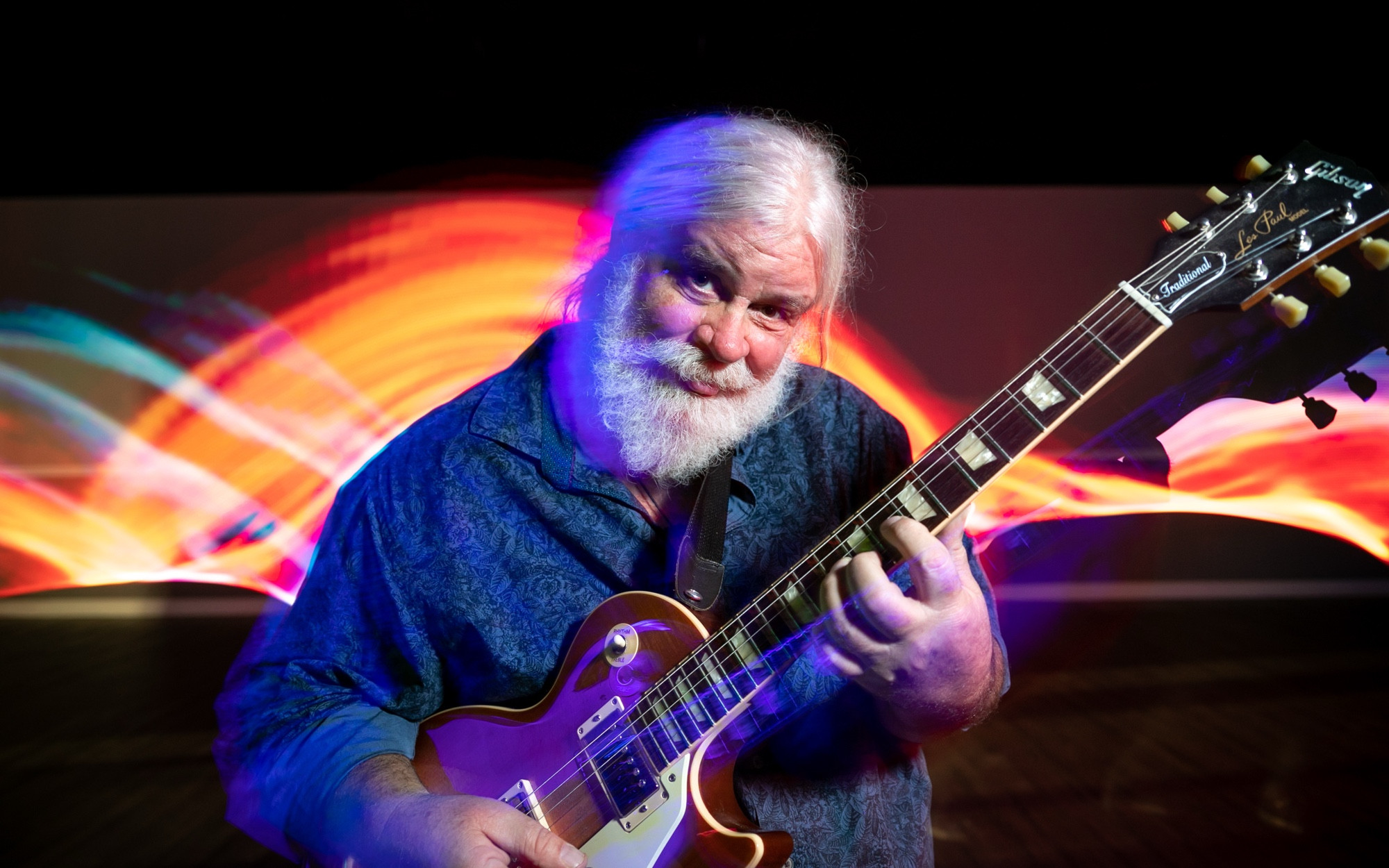 Photograph of Vince Herman playing guitar with rainbow spiral lights behind him.