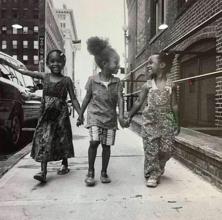 Monochrome photograph of three young Black girls walking g forward down a street holding hands and smiling