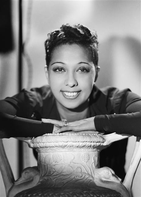 Monochrome photograph featuring a smiling Black woman with short hair wearing a dark top leaning on a pot and looking outwards 