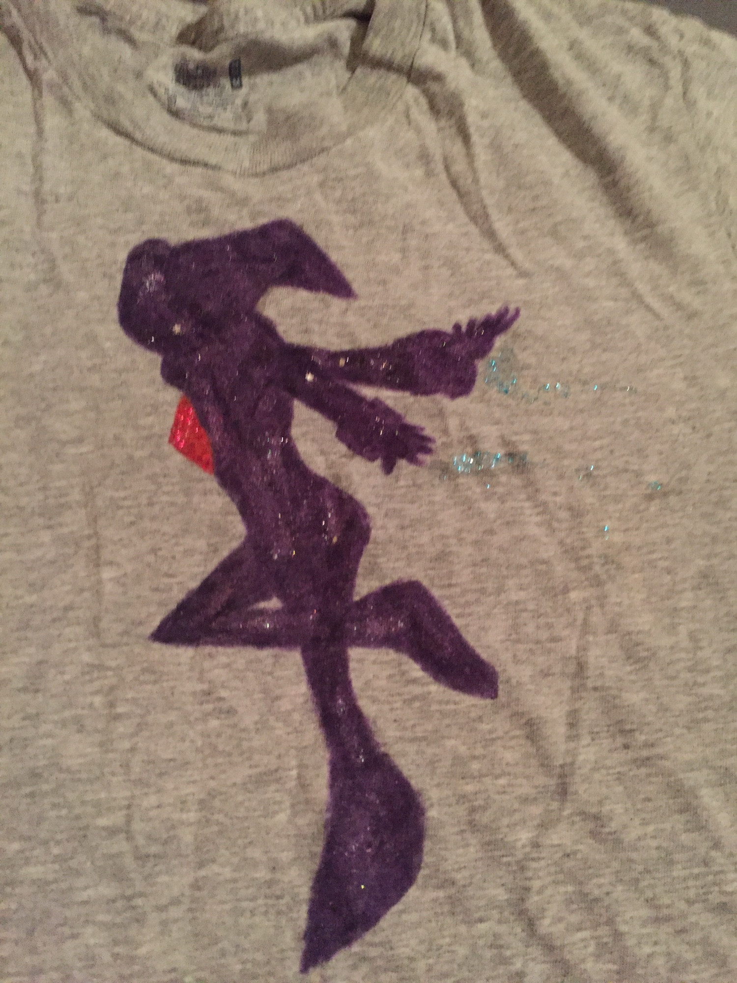 A simple silhouette drawn onto a gray t-shirt of NiGHTS. A jester-like character from the SEGA games: NiGHTS Into Dreams and NiGHTS: Journey of Dreams. The shirt seems to lean more into the NiD version.

There is glitter worked into the jewel, the shadow, and trailing from the hands. The blue glitter is meant to emulate the Star Trail (fanon term) or Twinkle Dust (JoD Term) that followed the character’s hands.

The design was made with fabric markers and nail polish. Lasting for years, funny enough. 