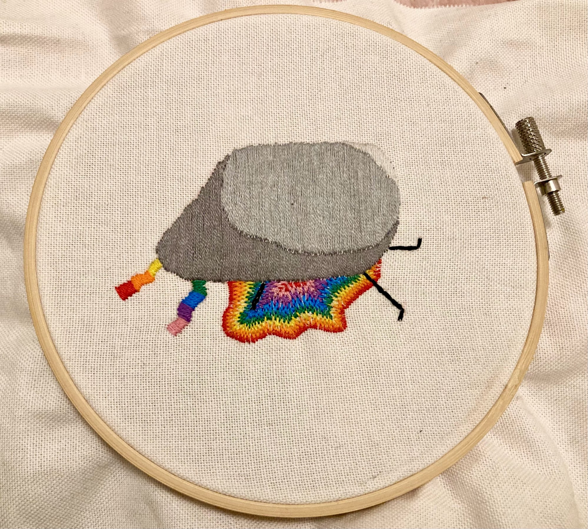 Embroidery of a stick figure of SpokeIsHere being crushed by a large rock. His legs and rainbow bandana stick out from under the rock and there is rainbow blood spilling out