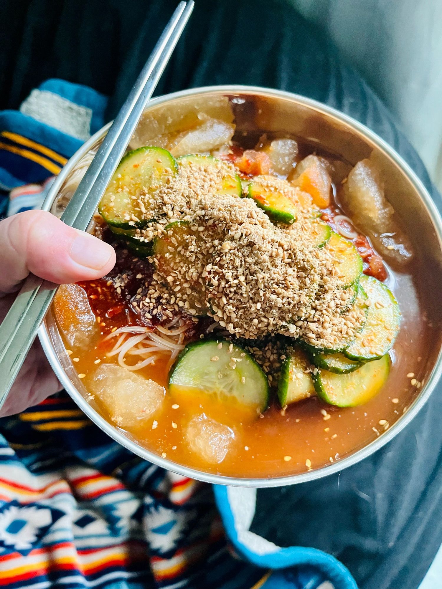 Bowl of Korean spicy cold noodles with cucumber salad and ground toasted sesame seeds in dongchimi broth.