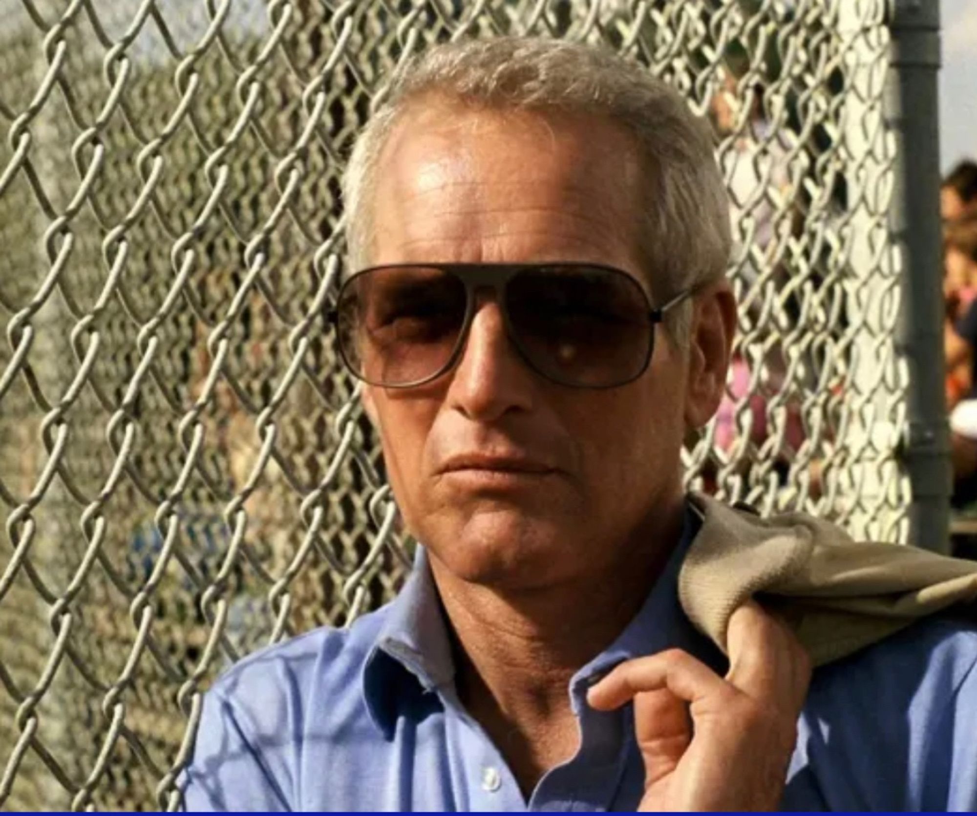 Paul Newman leaning against a fence looking cool as hell. He’s wearing sunglasses and a blue shirt and holding his jacket over his shoulder.