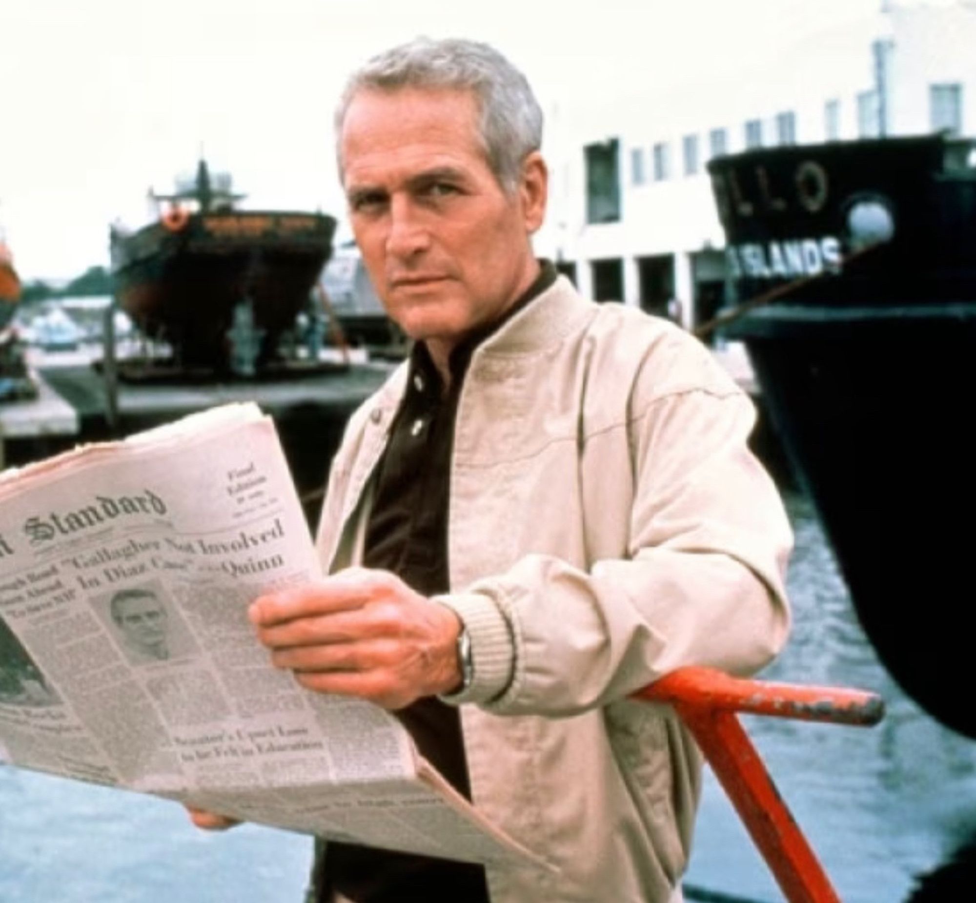 Paul Newman at a harbor or something leaning against a red metal thing looking cool as hell. He’s wearing a black shirt and a tan jacket and holding a newspaper.