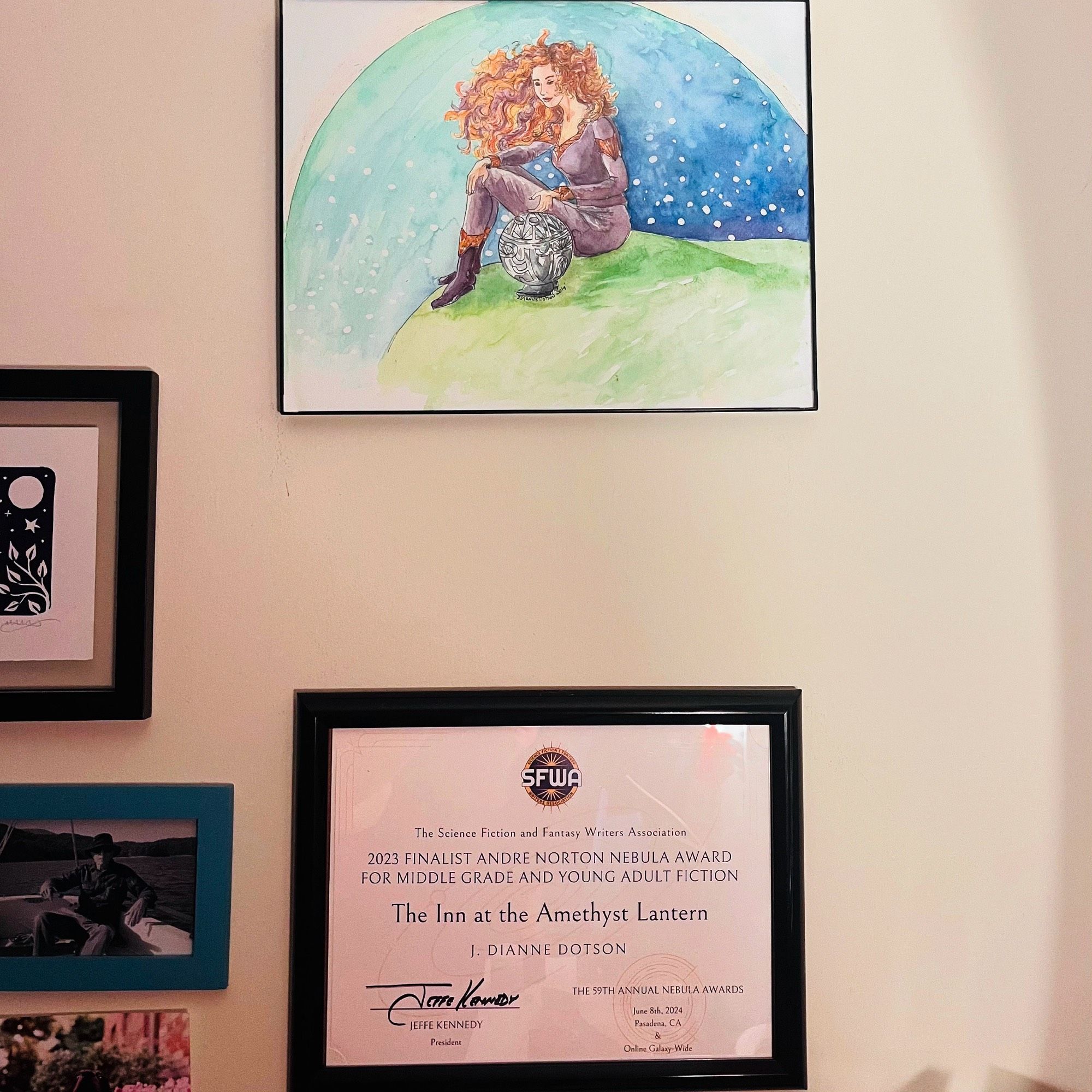 Framed pictures and certificate on a wall. The top picture is of the character Galla-Deia and the round robot Pliip, written and illustrated by Jendia Gammon from the Questrison saga.
The bottom frame holds the 2024 Nebula award finalist certificate for THE INN AT THE AMETHYST LANTERN written by Jendia Gammon under her J. Dianne Dotson pseudonym.