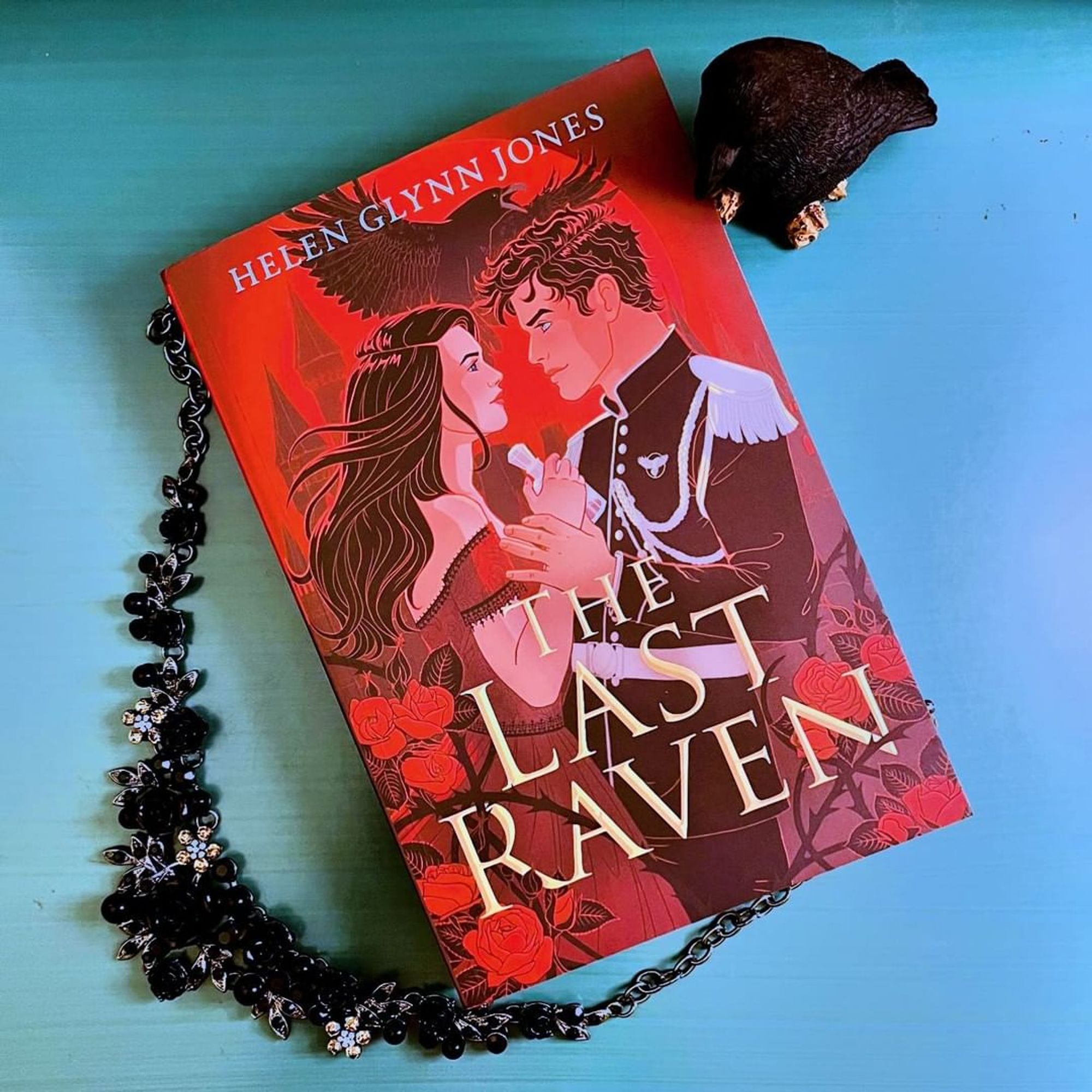 A book, The Last Raven by Helen Glynn Jones (with a young woman and young man facing each other, a raven flying overhead, and a red background), on a teal desk. A black floral jeweled necklace and a small raven statue surround the book.