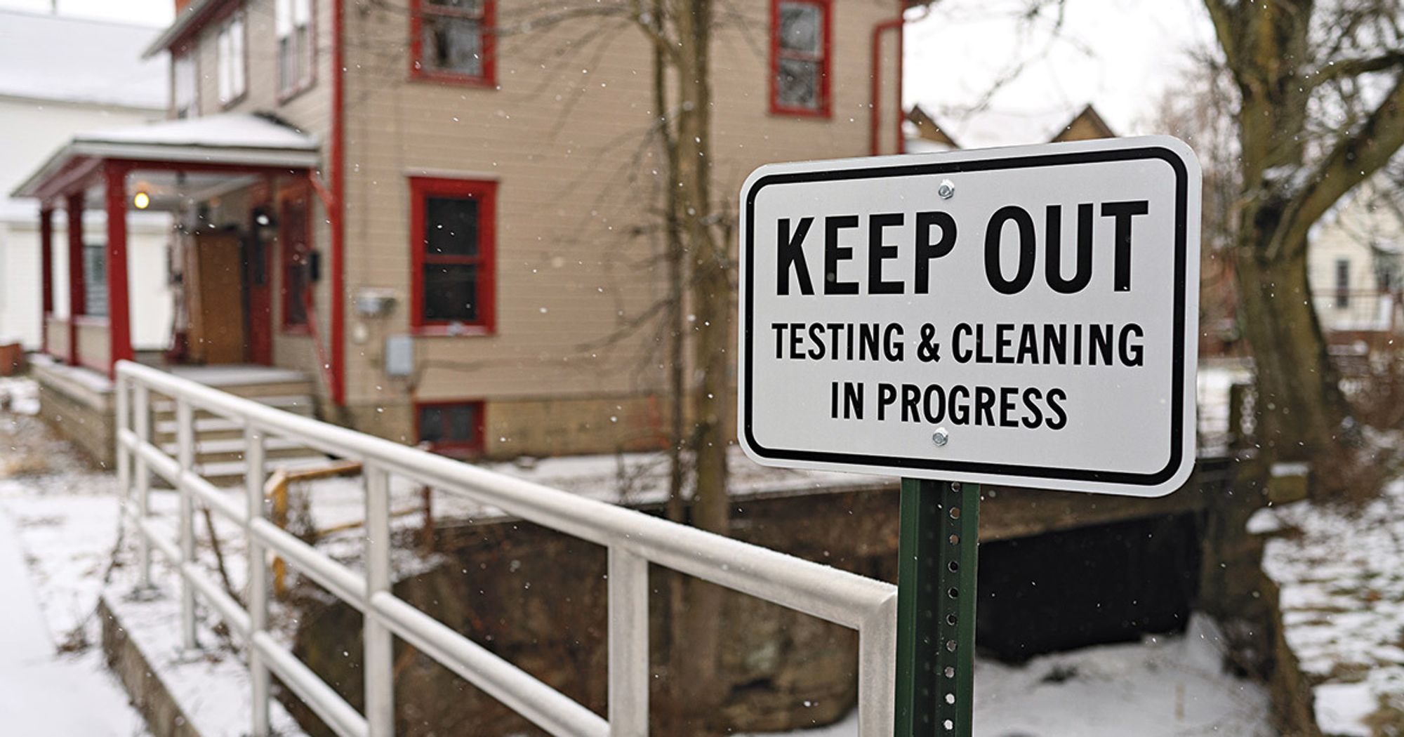 A "Keep Out; Testing & Cleaning in Progress" sign is in the foreground next to a tan house, snowflakes falling around it.