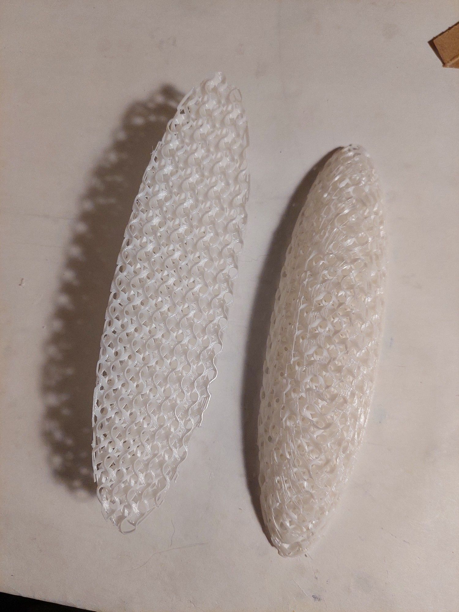 A poorly-lit photo of two simple oblong pads, 3d printed with a flexible white filament. They have no outer shells, so the infill makes up the entire structure.