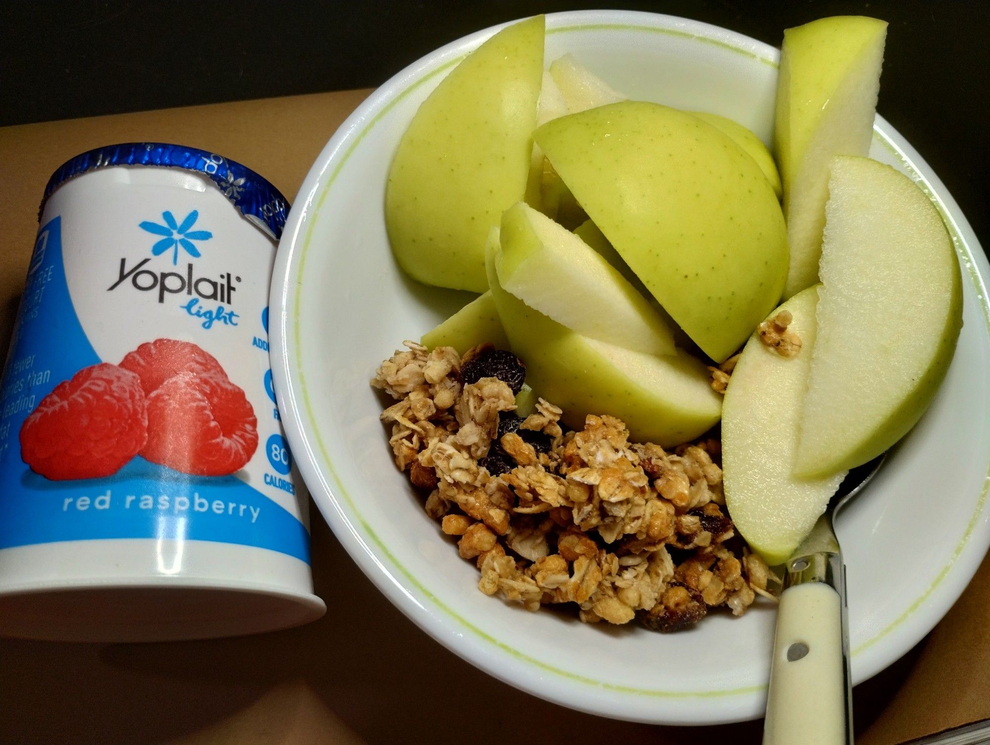 White bowl on the right containing a white handled spoon, a roughly cut up green apple and granola. To the left there is a single serving container of yoplait light red raspberry yogurt. The yogurt pot is white with blue branding text and a faded image of three red raspberries.