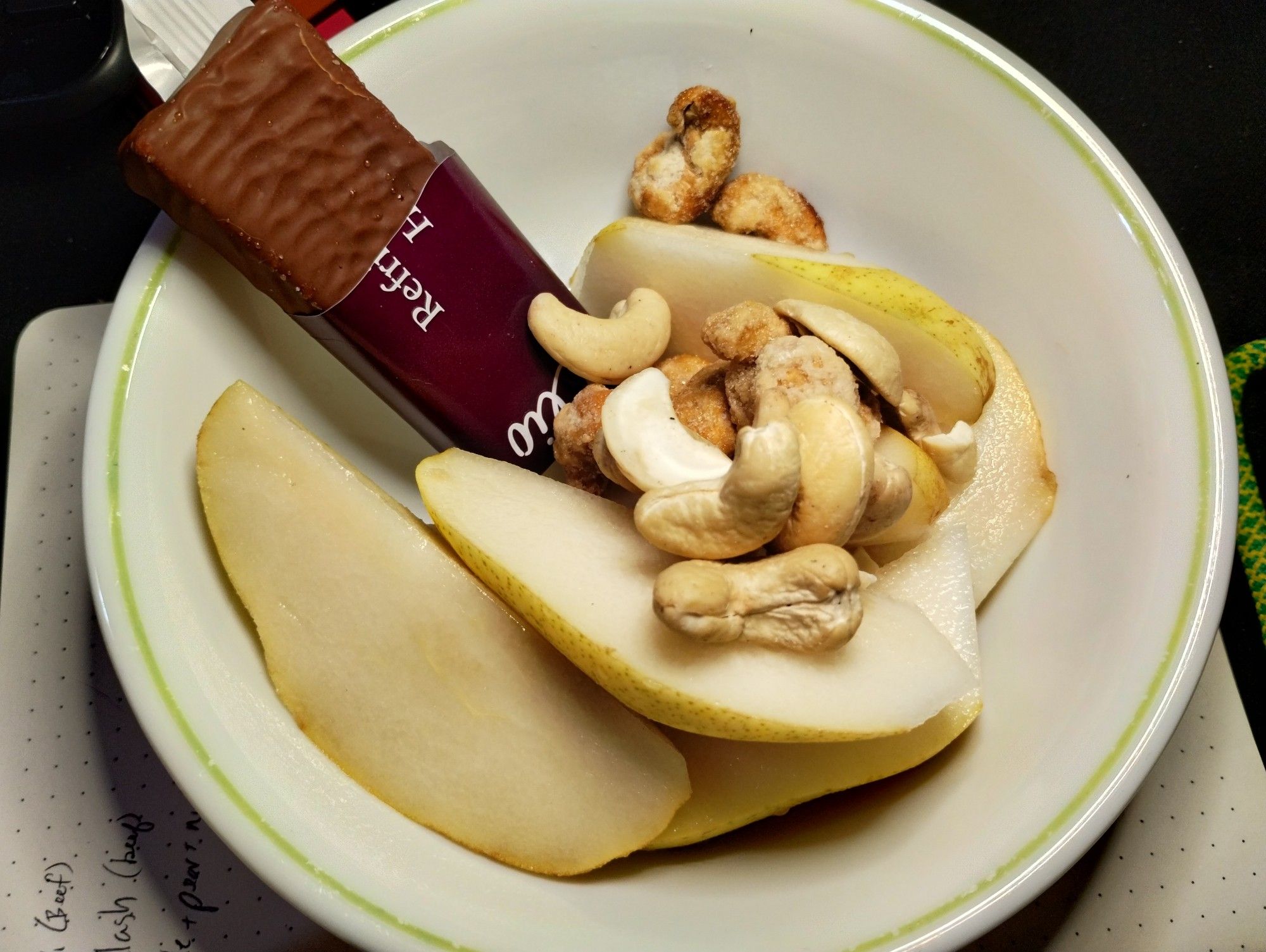 White bowl with a green stripe edge. Inside the bowl is a half unwrapped bar coated in chocolate, the wrapper is brown. There is a sliced up pear. There are two kinds of cashew, one pale and the other kind with a brown crunch coating